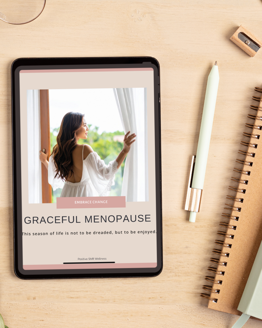 A tablet displaying an article about embracing change during graceful menopause, with a woman looking out a window on the screen. The tablet is placed on a light wooden desk alongside a white pen, a small wooden pencil sharpener, and a spiral-bound notebook.