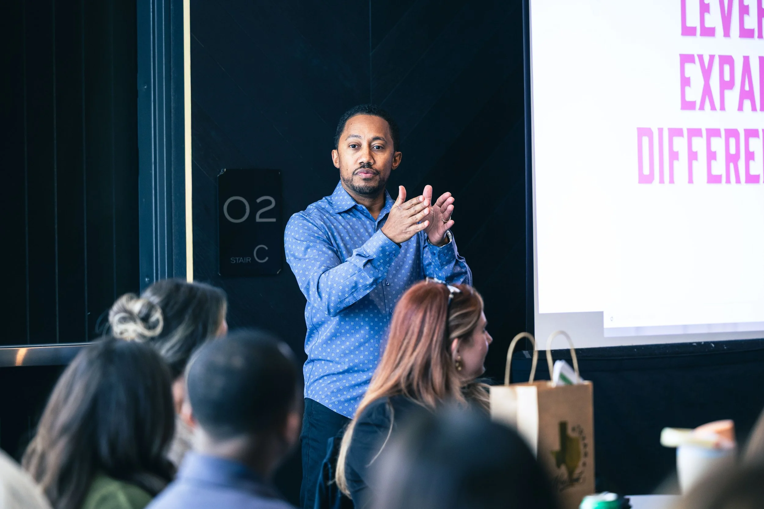 Man in blue shirt standing in front of a presentation screen, clapping while an audience listens.