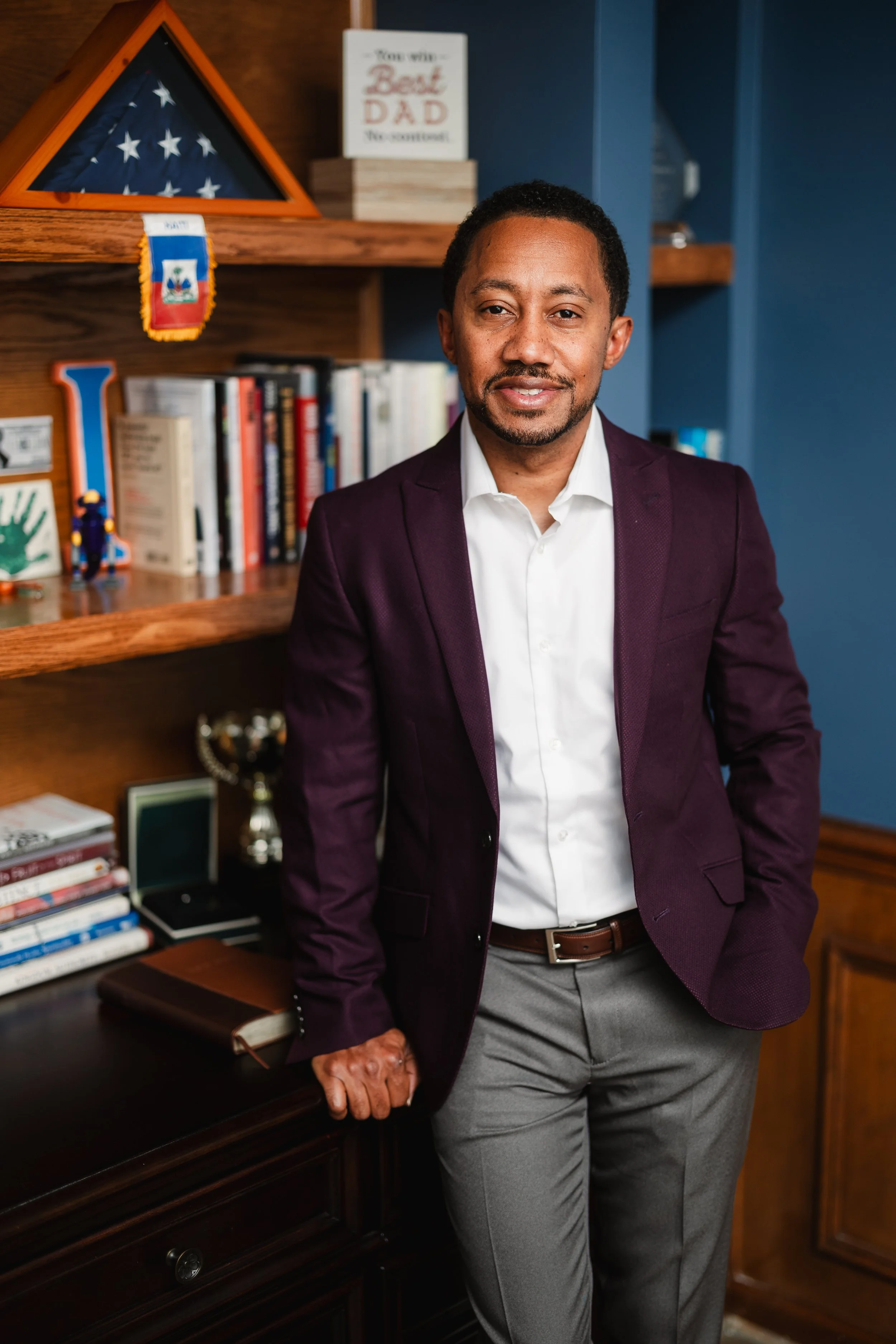 A man wearing a dark purple blazer, white shirt, and gray pants standing next to a wooden desk with his right hand on it. Behind him, there is a bookshelf filled with books, a framed American flag, and a framed sign that says "You win some, Best Dad, You win some." The background wall is painted dark blue.