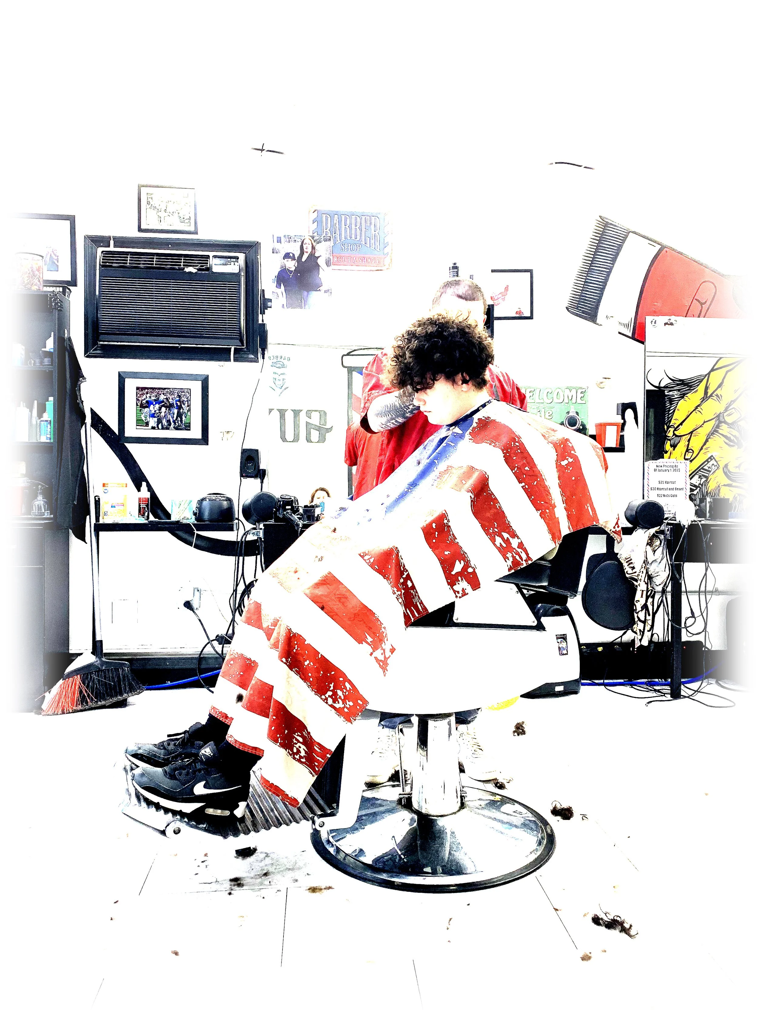 A person with curly hair getting a haircut at a barbershop. The barber is standing behind them. The person is sitting in a chair covered with a red, white, and blue striped barber cape. The barbershop has various framed pictures, a sign that says "Barber", and equipment on the counters.