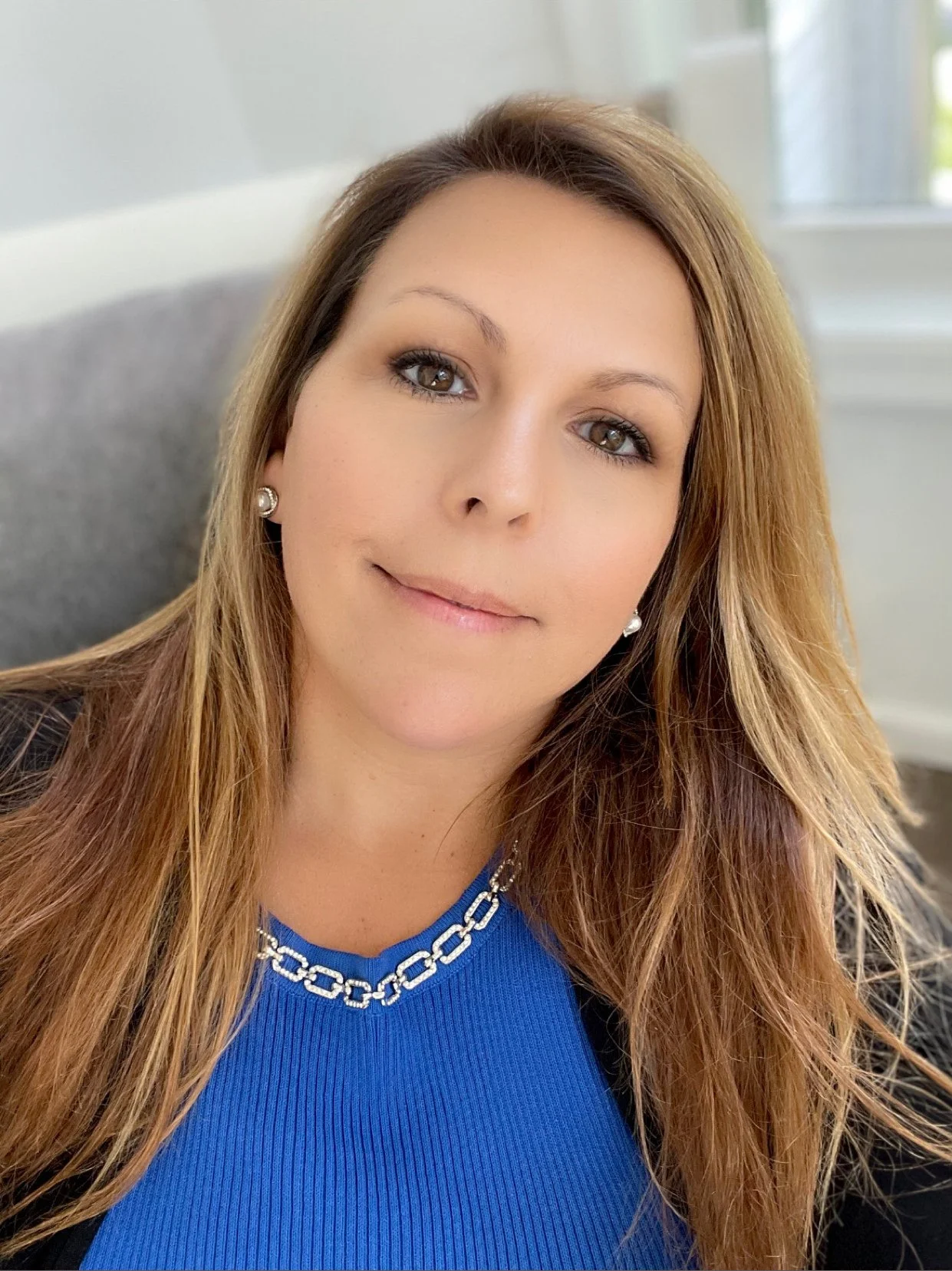 A woman with long, light brown hair, wearing a blue top with a silver chain necklace, smiling softly with a neutral background and window in the background.