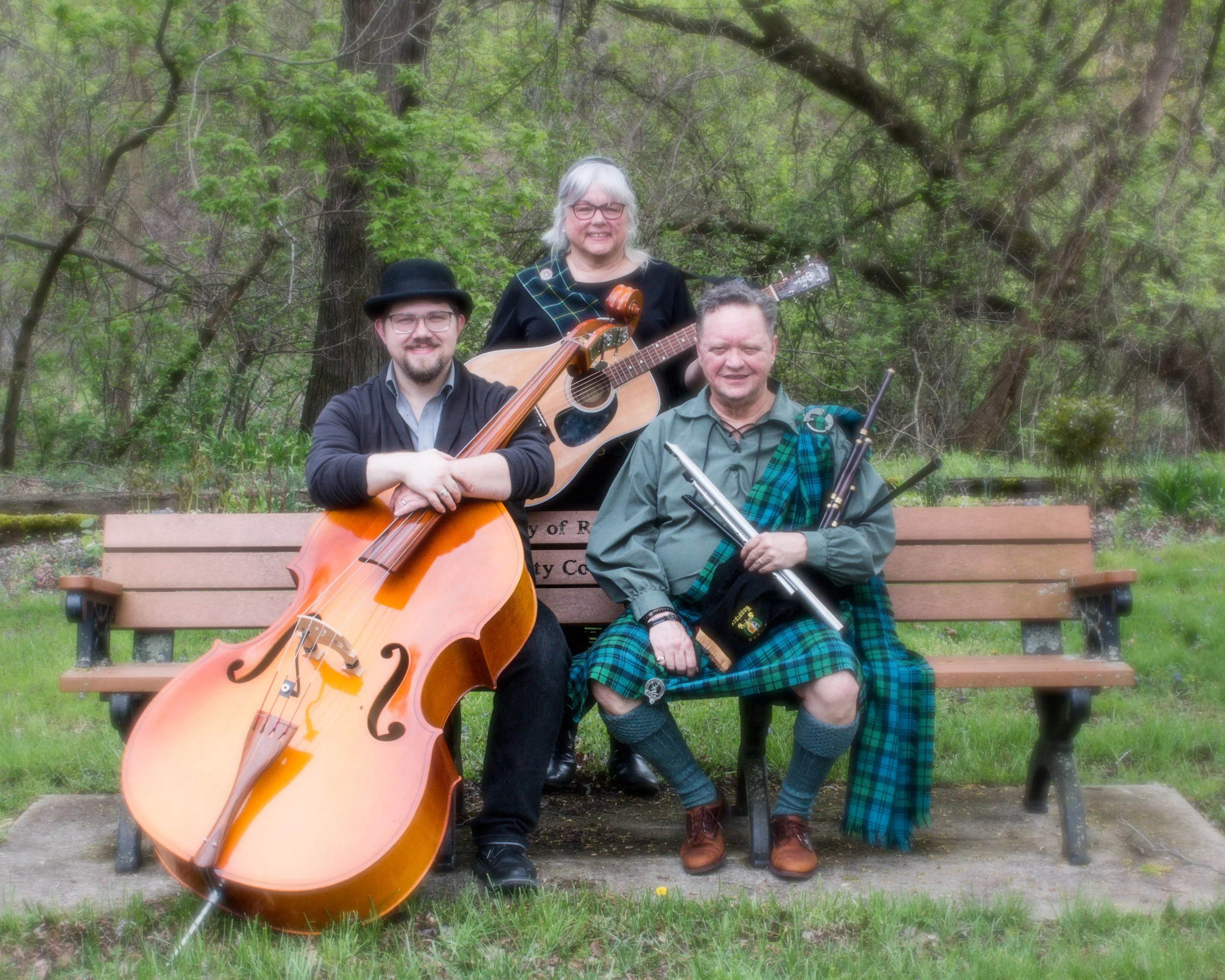 Three people sitting on a park bench with musical instruments, surrounded by trees and greenery. One person with glasses holds a double bass, another with a guitar, and the third person, dressed in traditional Scottish attire, holds bagpipes.