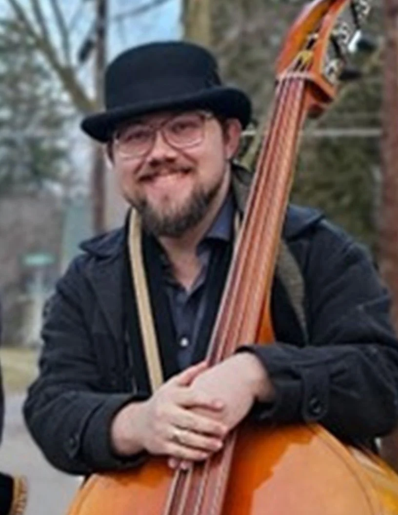 A man with glasses, a beard, and a mustache, wearing a black top hat and black jacket, is smiling and holding a double bass outdoors with trees in the background.