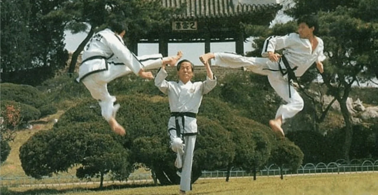 Three people practicing martial arts outdoors in traditional uniforms, performing flying kicks and high moves near a large tree and a traditional building.
