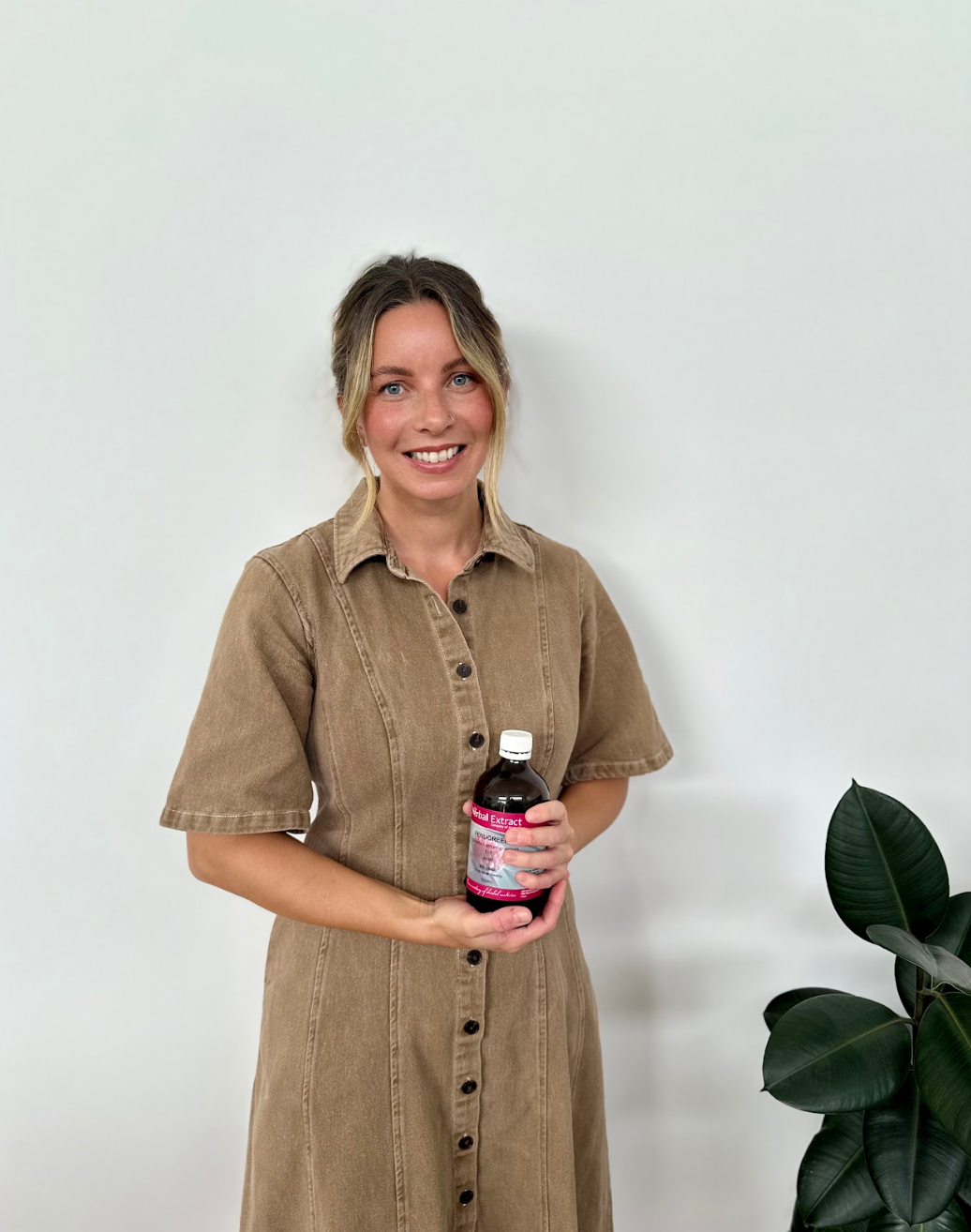 A woman with blonde hair, wearing a brown button-up dress, standing against a white wall, holding a bottle of herbal extract, next to a large green plant.