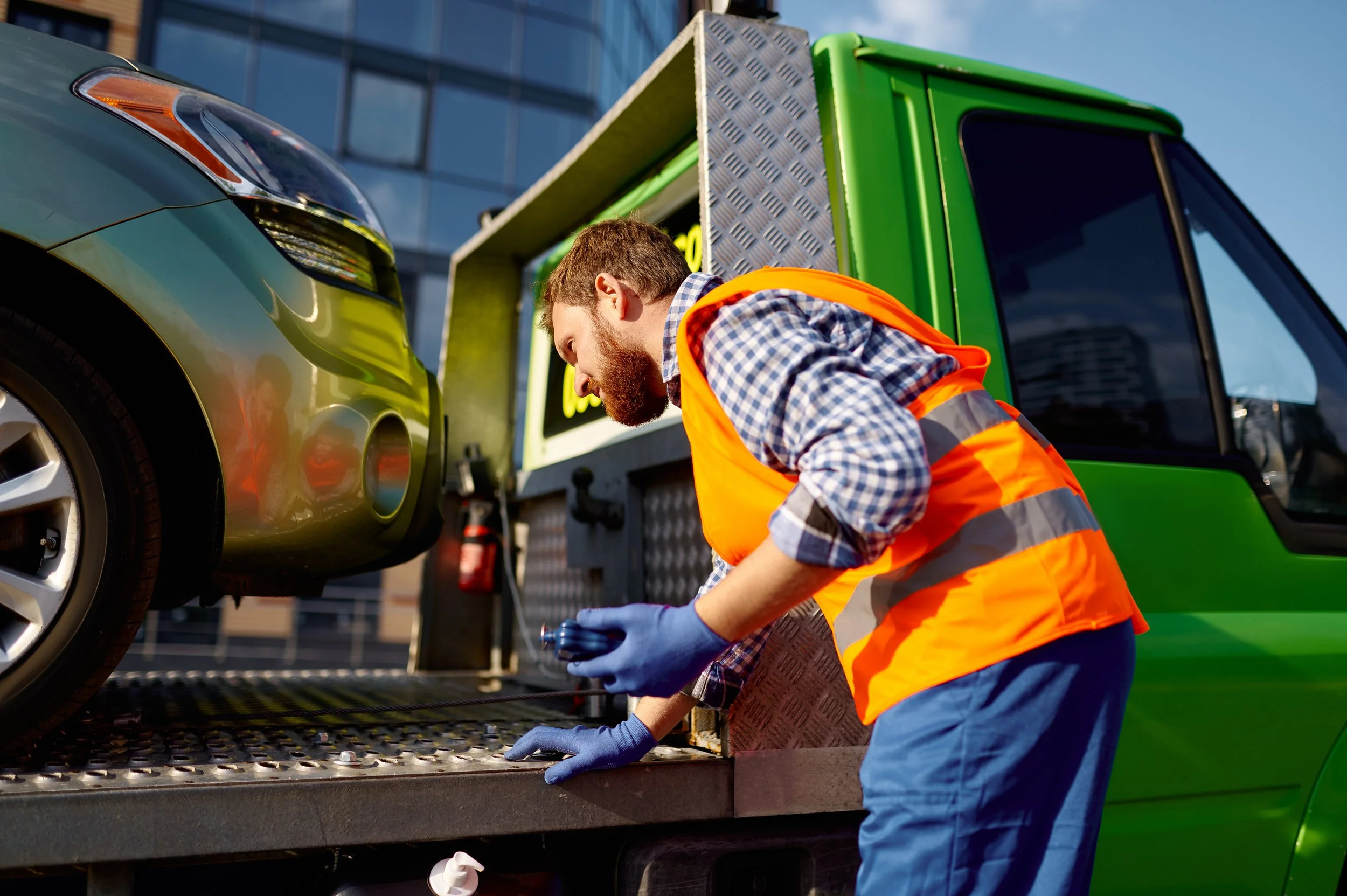 A tow truck operator in an orange safety vest and blue gloves inspecting a green car on a flatbed tow truck.