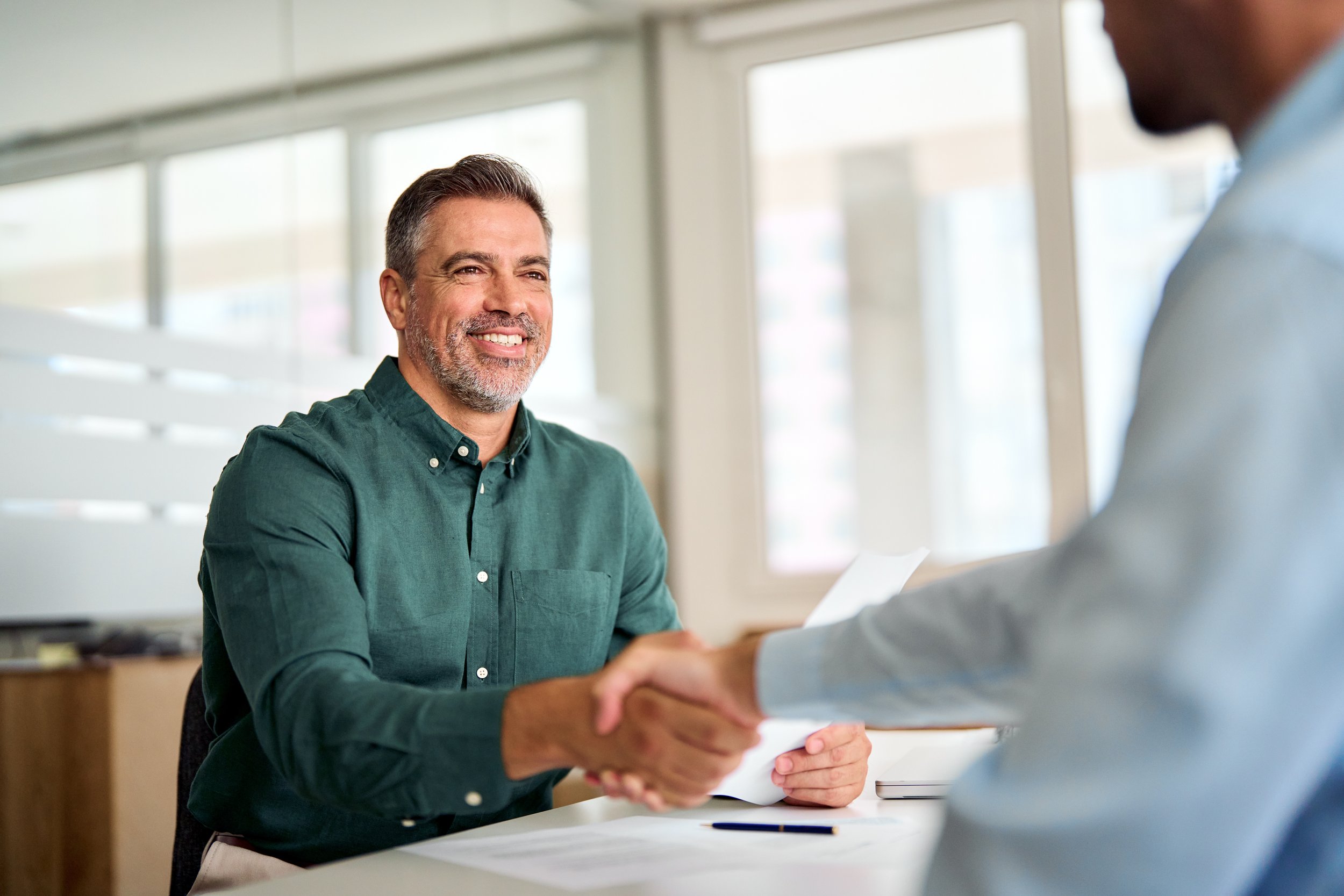 Two men shaking hands during a job interview or business meeting in an office setting.