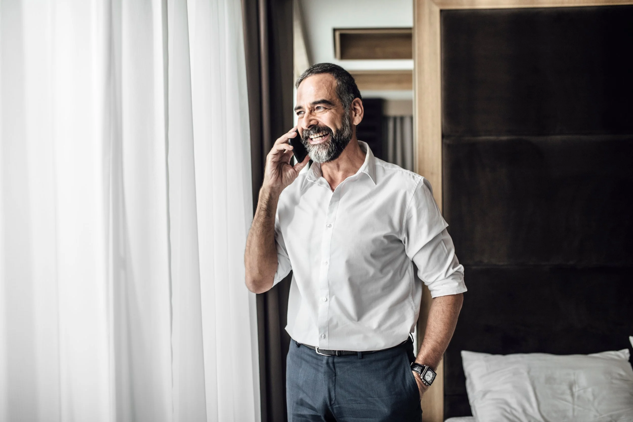A smiling man with a beard, dressed in a white shirt and gray pants, talking on a phone inside a hotel room.