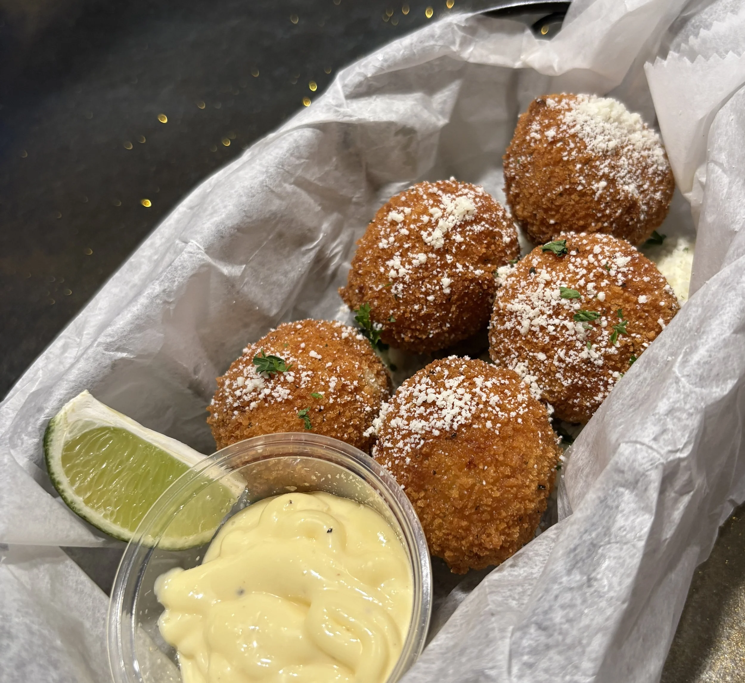 Six golden-brown croquettes topped with grated cheese and herbs, served with a lime wedge and a small container of creamy yellow dipping sauce.