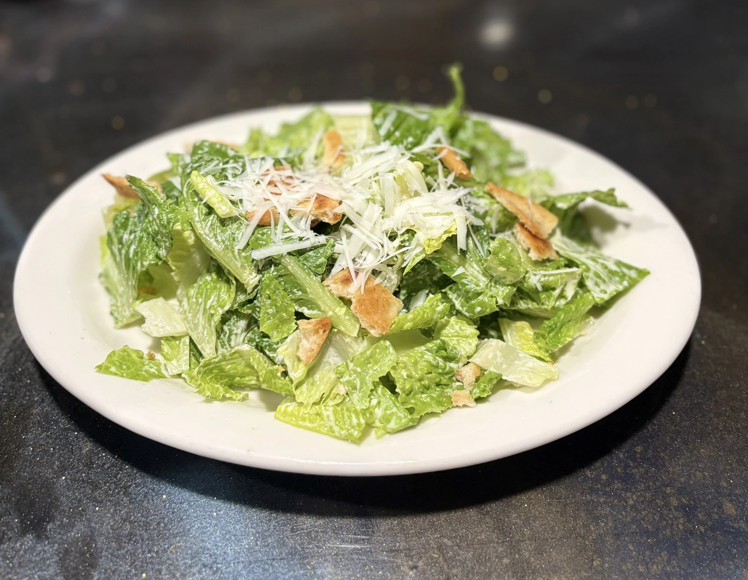 A plate of Caesar salad with Romaine lettuce, grated cheese, and croutons on a dark surface.
