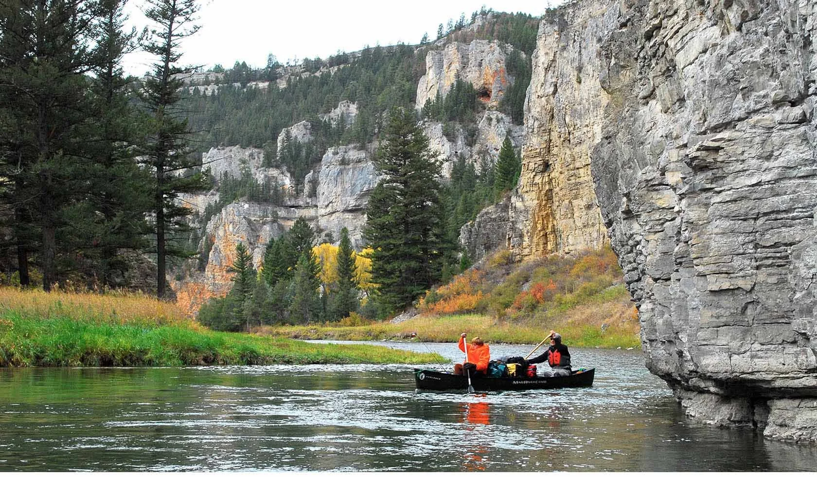Two people in a black canoe paddling on a river surrounded by tall rocky cliffs and forested mountains.