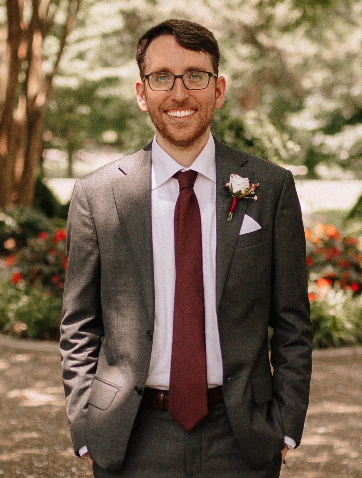 A smiling man in a gray suit with a white shirt, burgundy tie, and glasses, standing outdoors with a background of trees and flowers.