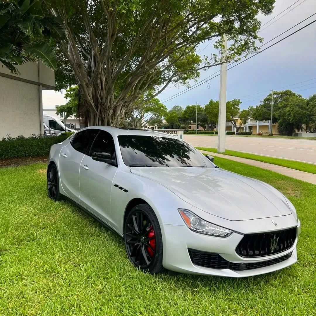 A silver Maserati sports car parked on a grassy area next to a sidewalk and a large tree, with a street and buildings in the background.