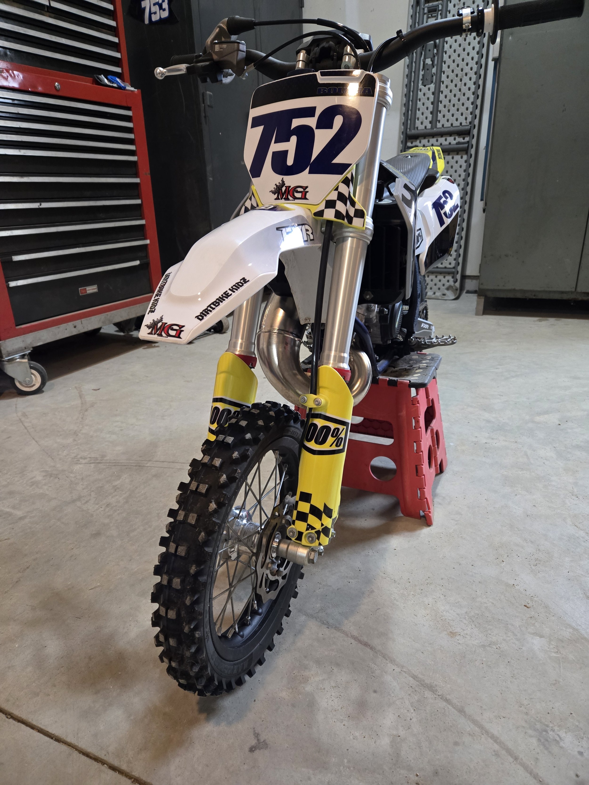 A dirt bike with No. 152, racing number, parked in a garage on a red stand, with front tire and suspension visible, and various tools and storage cabinets in the background.