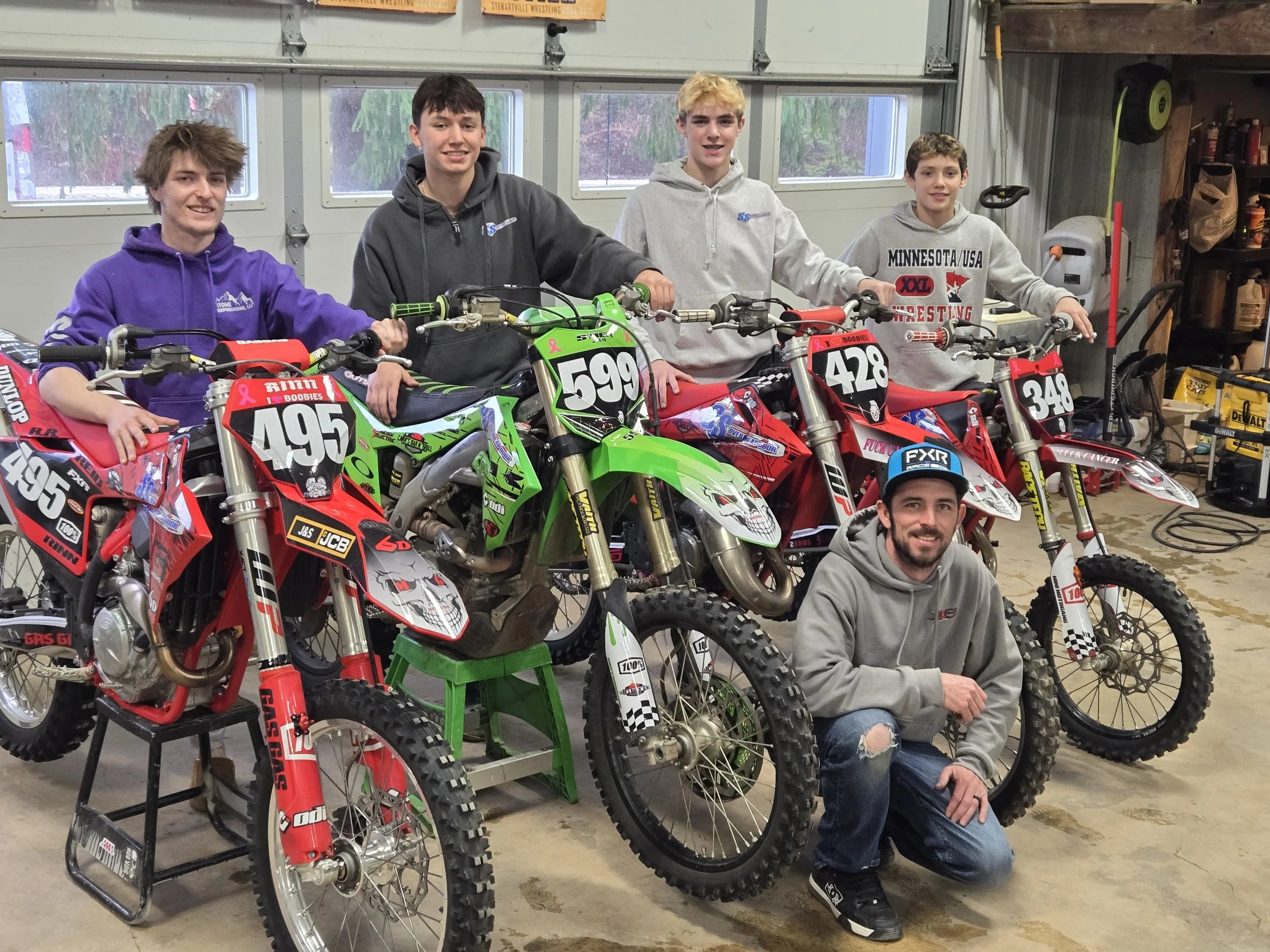Group of five young men in a garage with four dirt bikes, some sitting on them and one kneeling in front, all smiling.