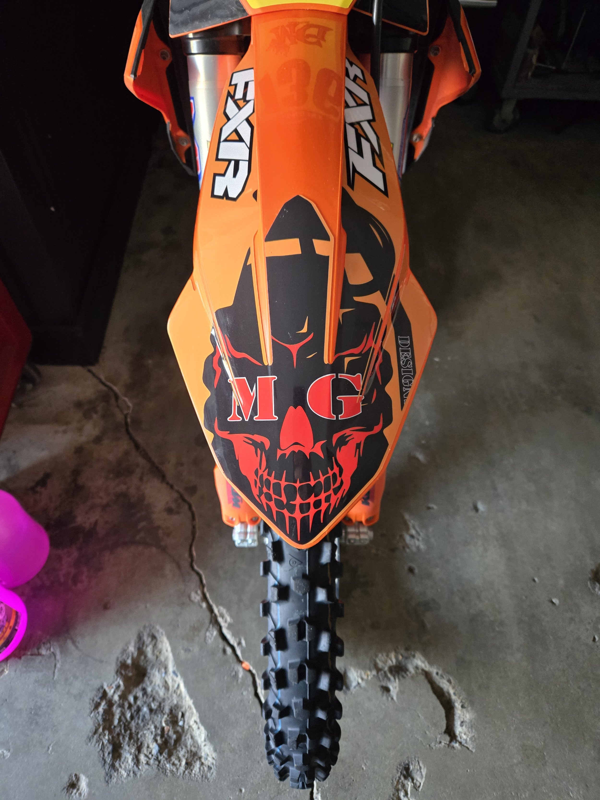 Top view of an orange motocross bike with a skull decal on the front fender, featuring red and black details and the letters 'MG' incorporated into the design.