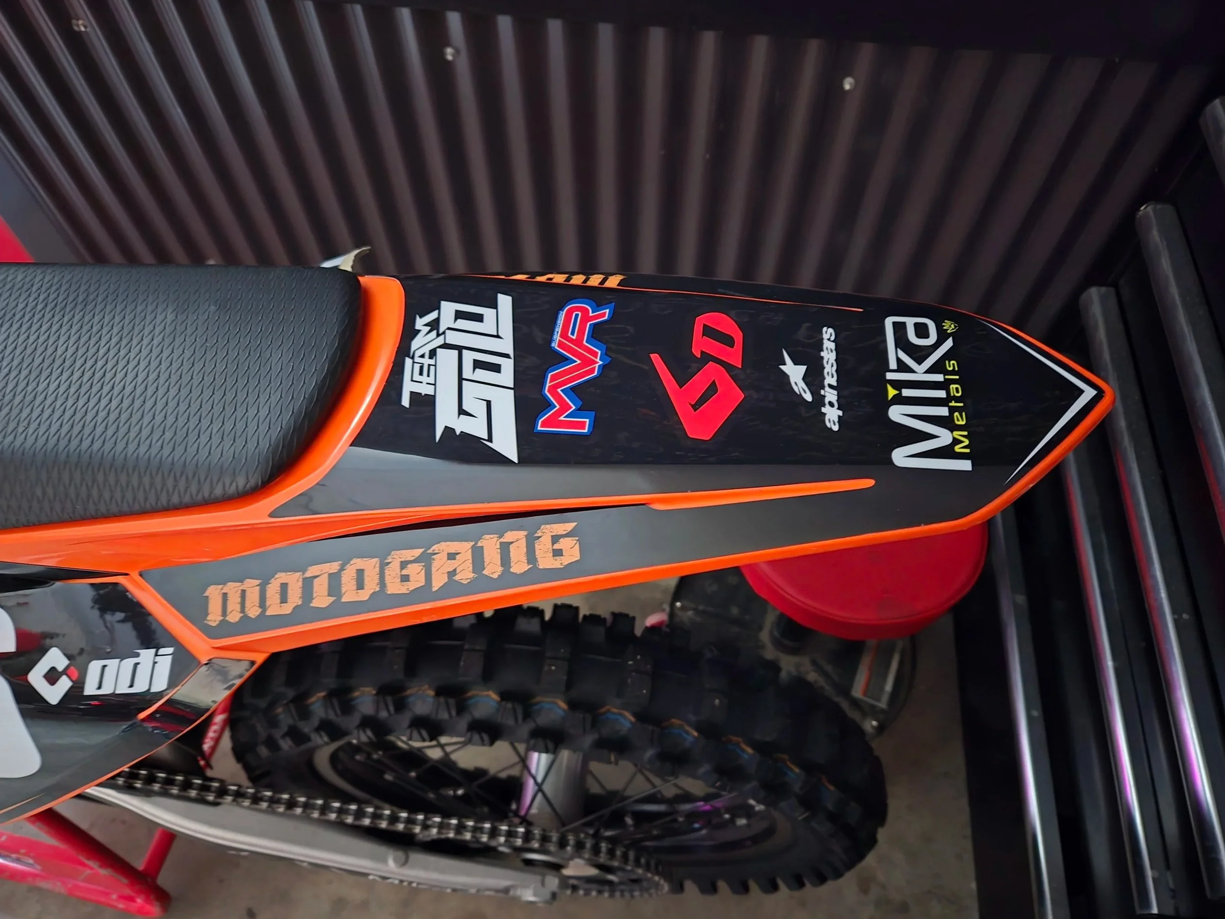 Close-up of a dirt bike's side panel with various sponsorship stickers, including Team Gold, MVR, SD, Alpinestars, and Mika Metals, featuring a black and orange color scheme.