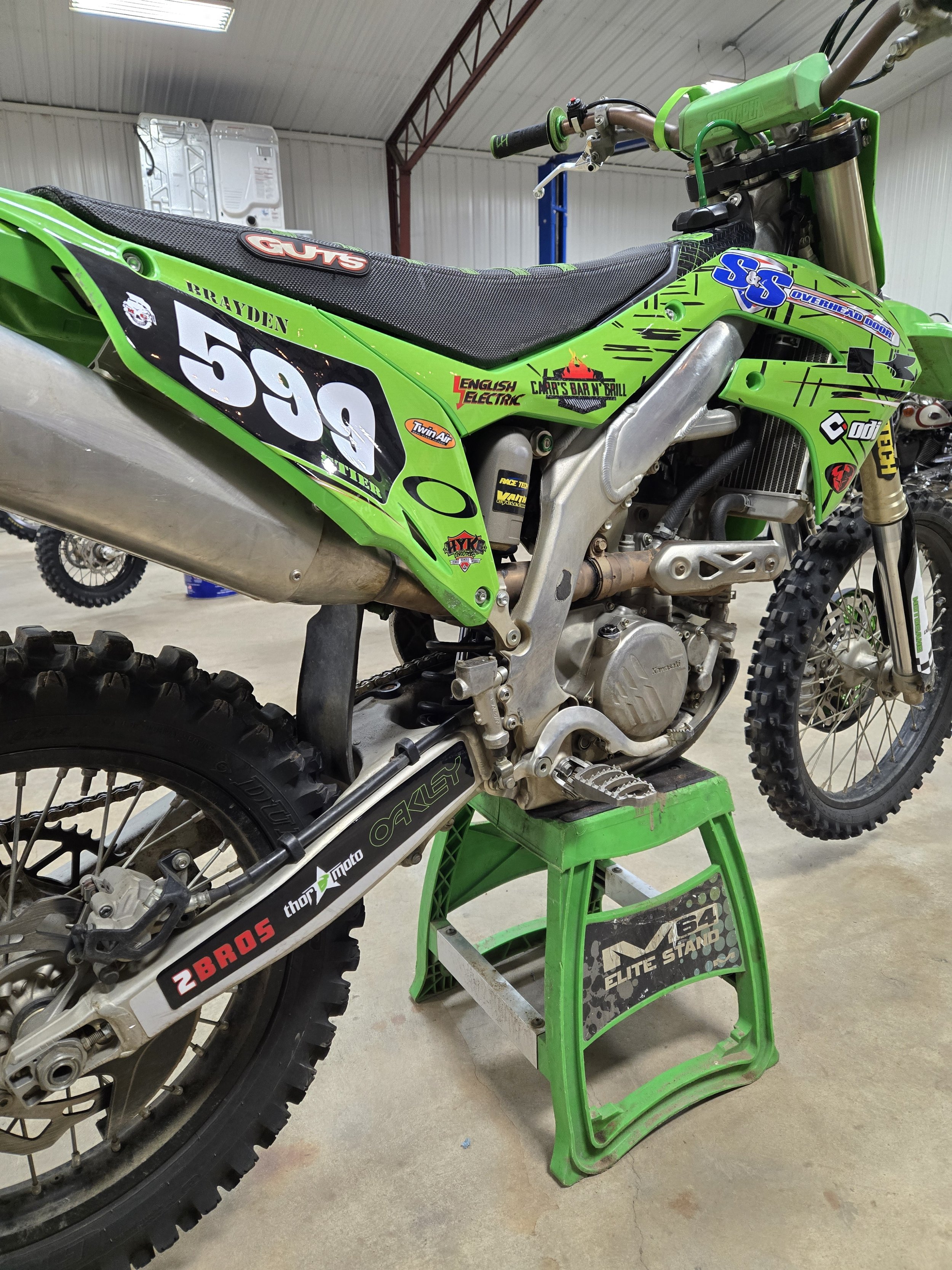 Green motocross bike on a green stand inside a garage, with various stickers and decals on it.