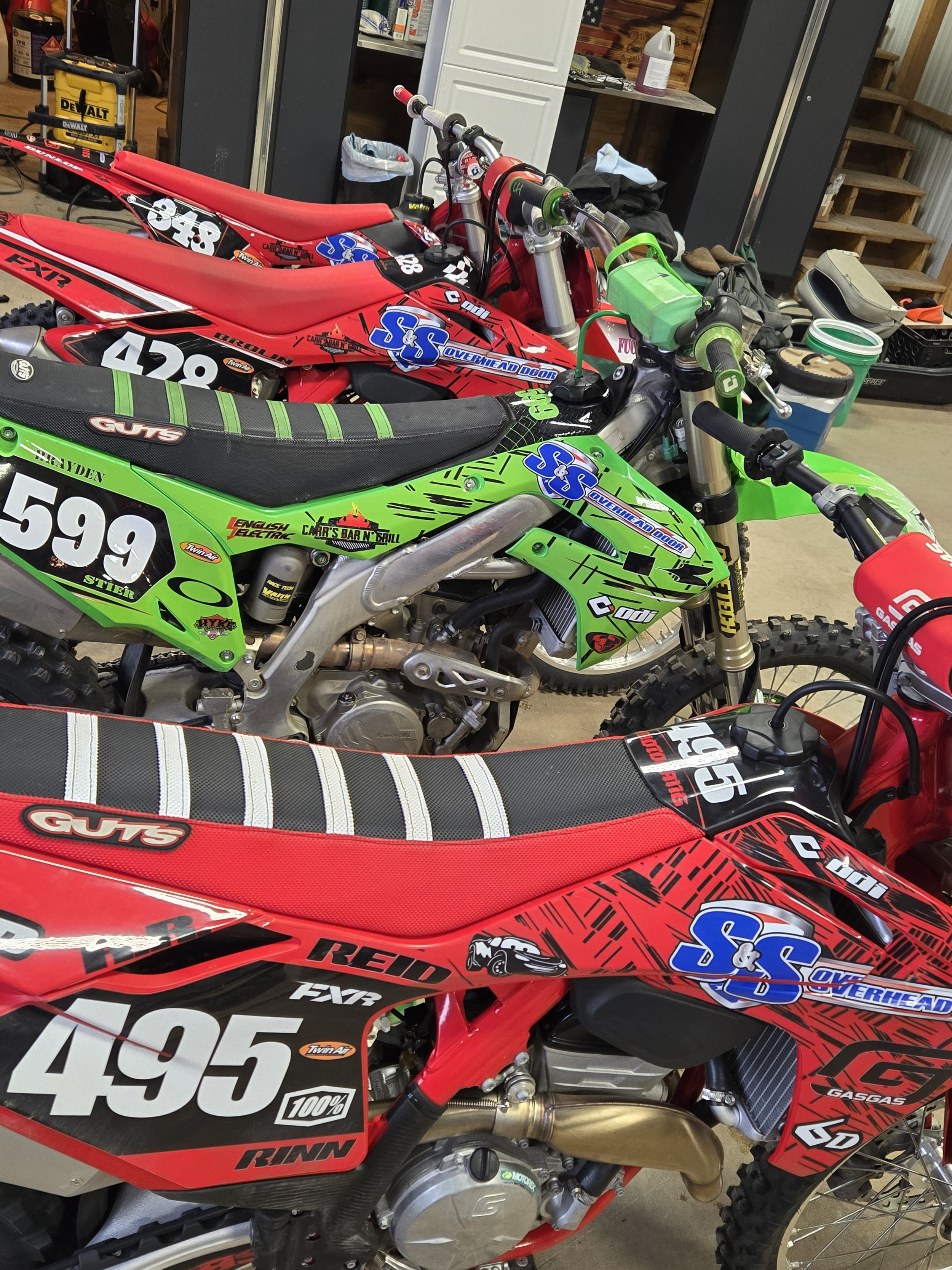 Several motocross bikes with racing numbers and decals inside a workshop with tools and storage shelves.