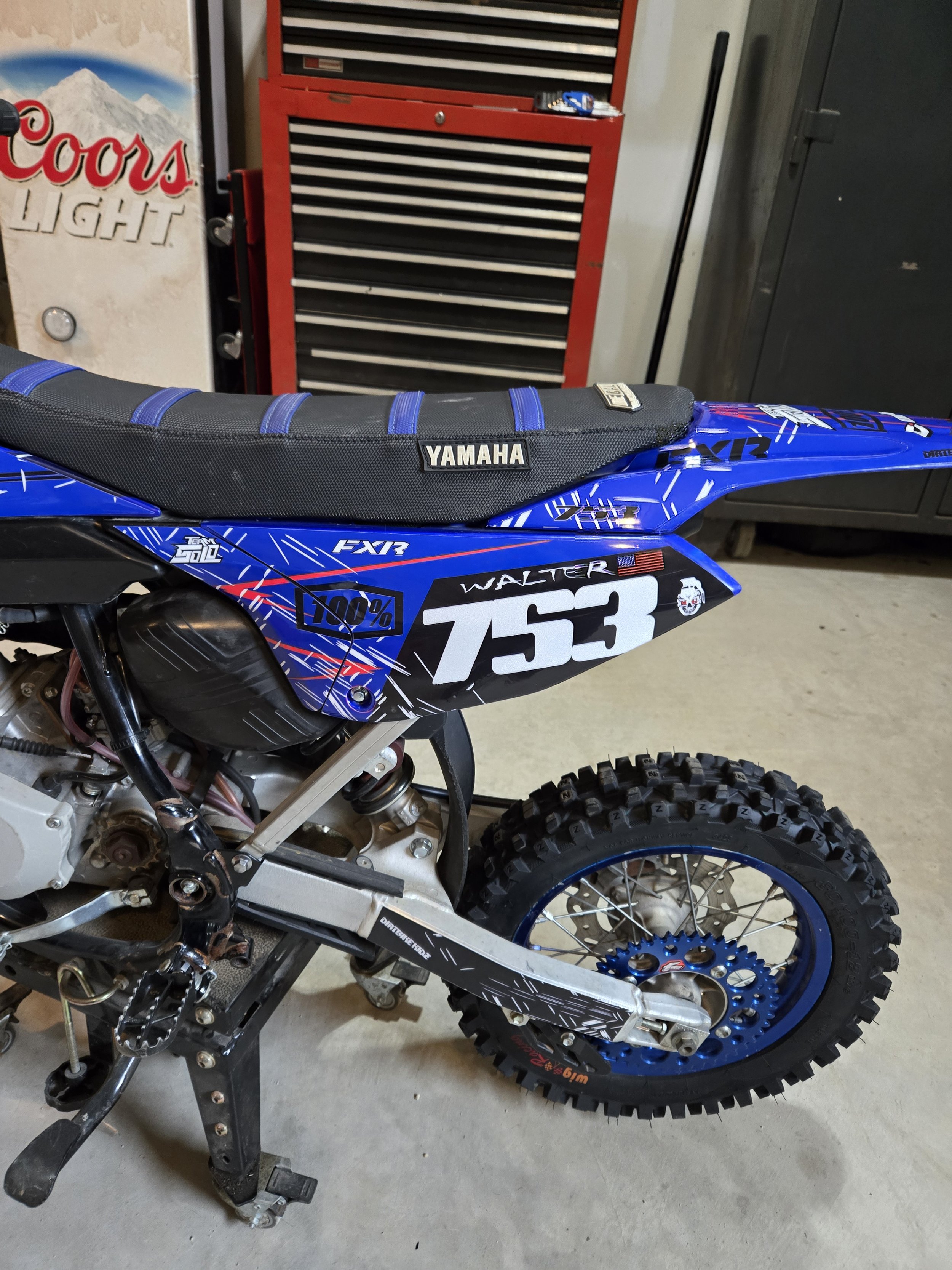 A Yamaha dirt bike with the number 753, parked in a garage with a red tool chest and a Coca-Cola light display in the background.