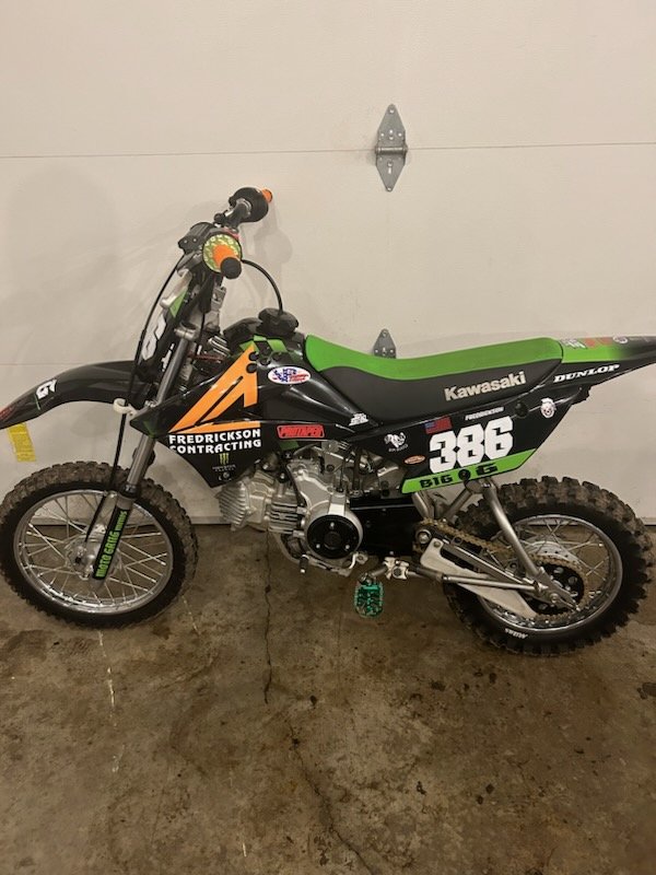 Green and black Kawasaki dirt bike with racing stickers, leaning against a white garage door on a concrete floor.