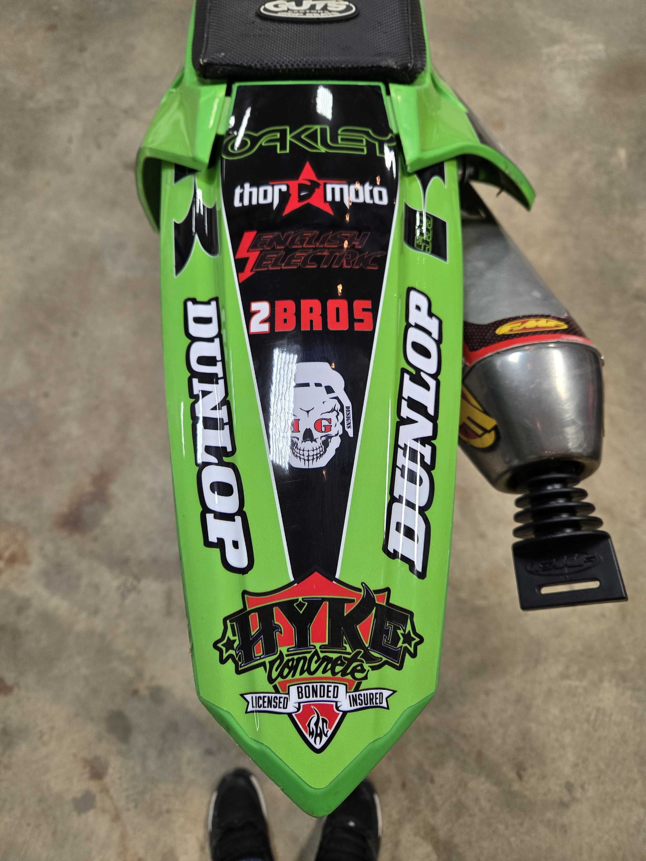 Top view of a green dirt bike with various stickers, including brands and logos, on the front fender.