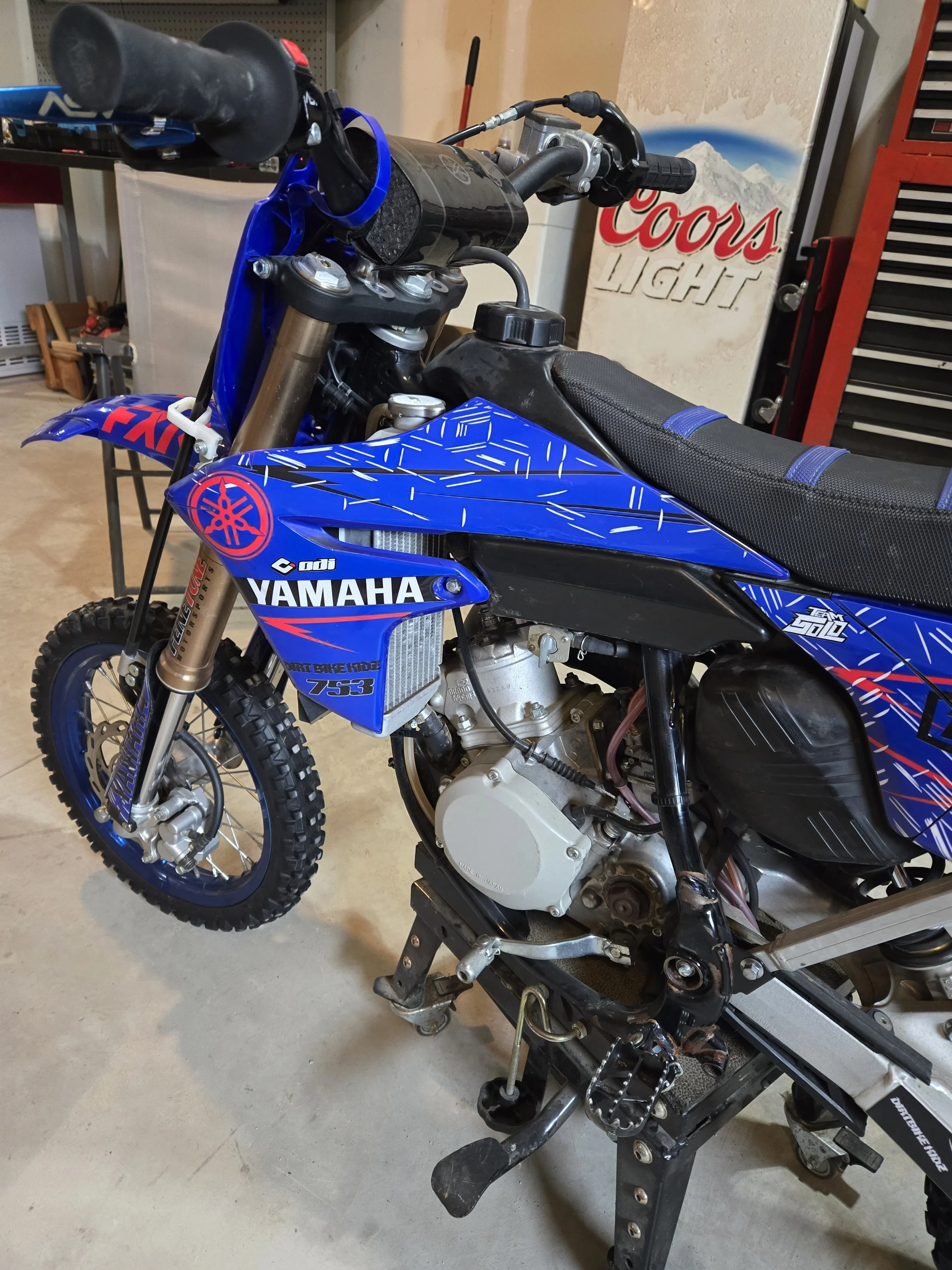 A partially assembled Yamaha dirt bike with blue and black bodywork, off-road tires, and a visible engine, inside a garage with tools and a Coors Light sign in the background.