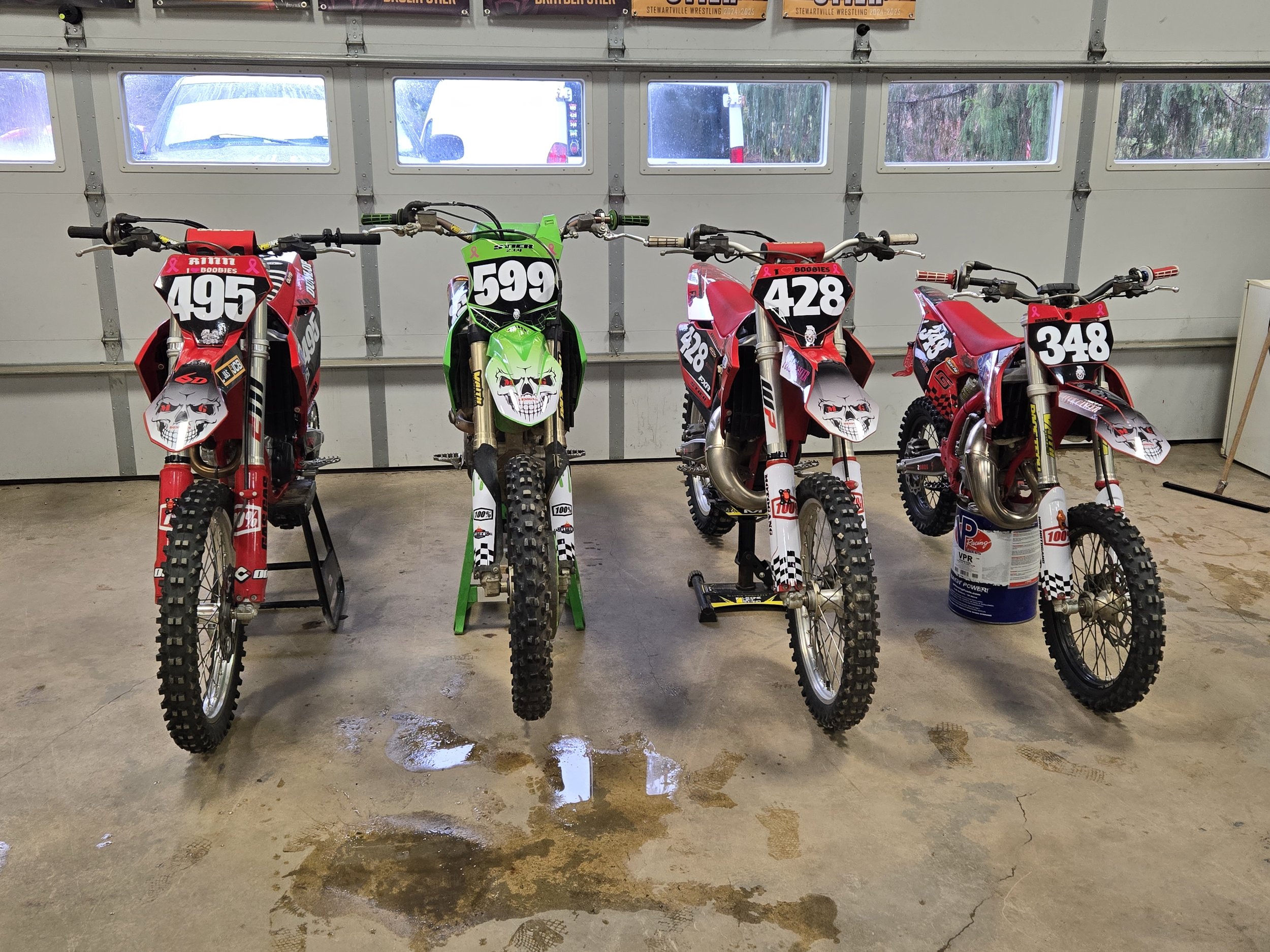 Four dirt bikes lined up in a garage, each with racing numbers 495, 599, 428, and 348, with decals and graphics, leaning on stands or resting on the floor, in front of a garage door with windows showing the outdoors.