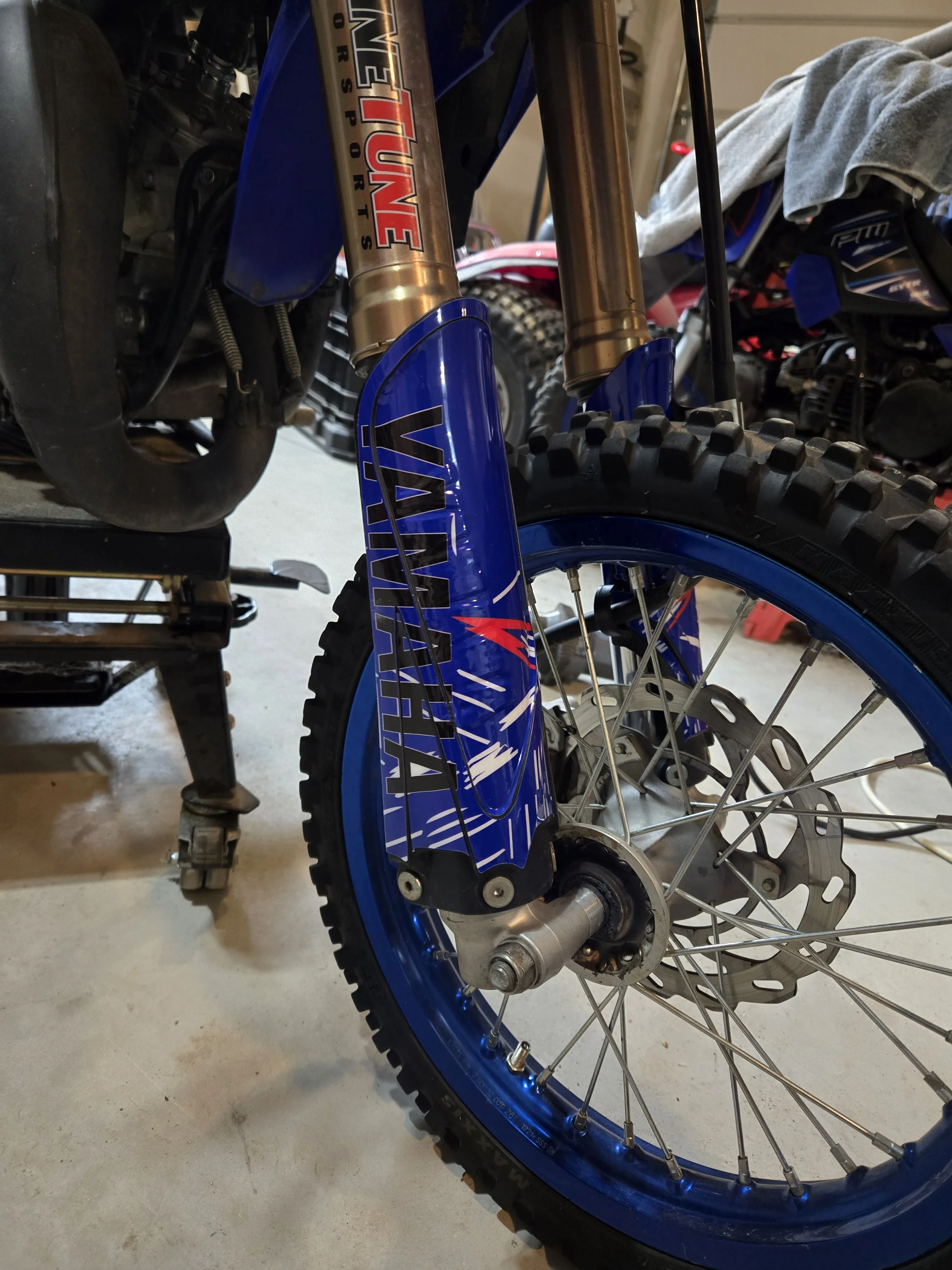 Close-up of a Yamaha off-road motorcycle's front wheel and suspension with blue fender, disc brake, and knobby tire in a garage.