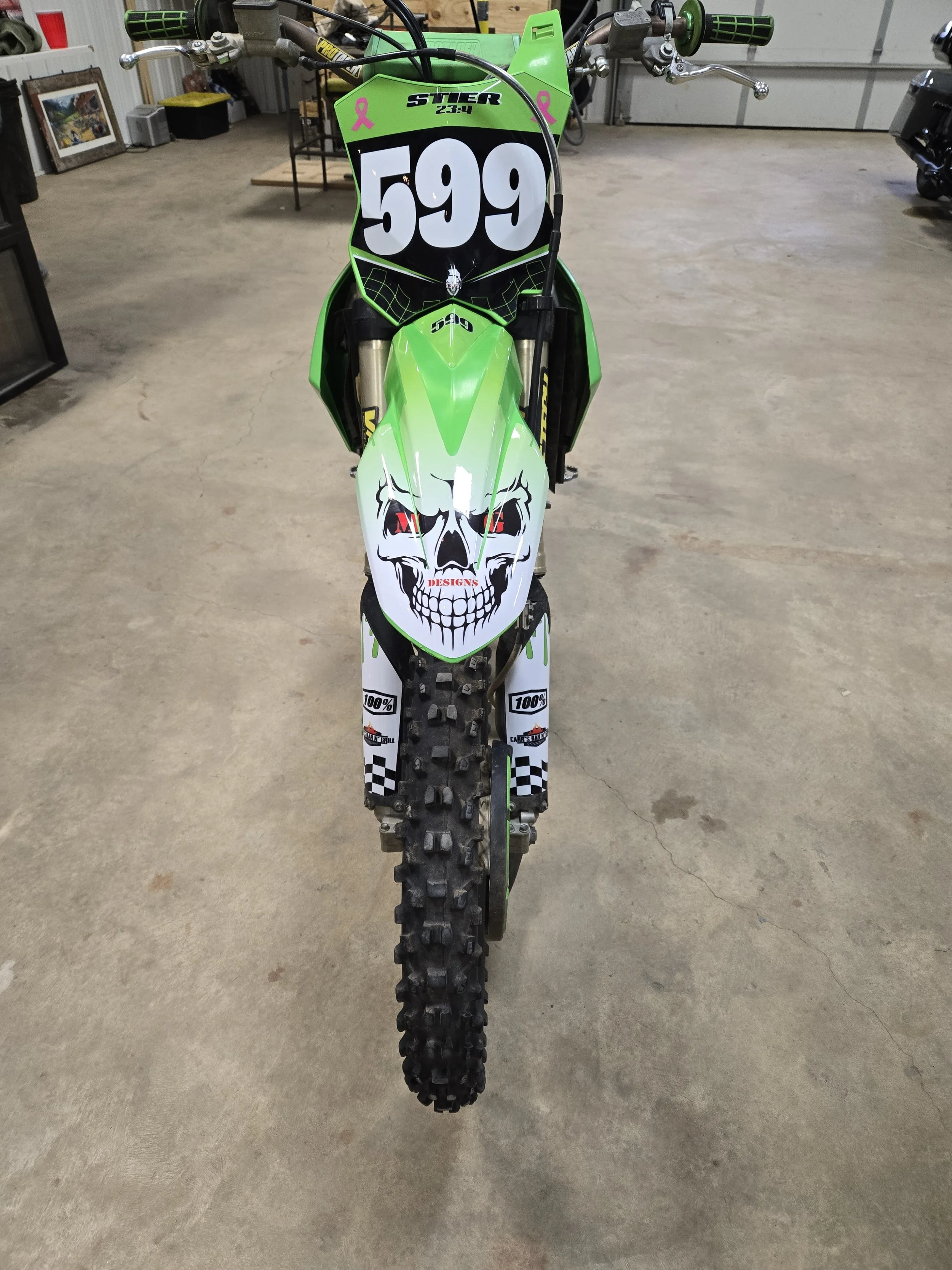 Green and white dirt bike with skull graphic on the front fender, racing number 599, and pink ribbon decals on the windshield, parked indoors on concrete floor.