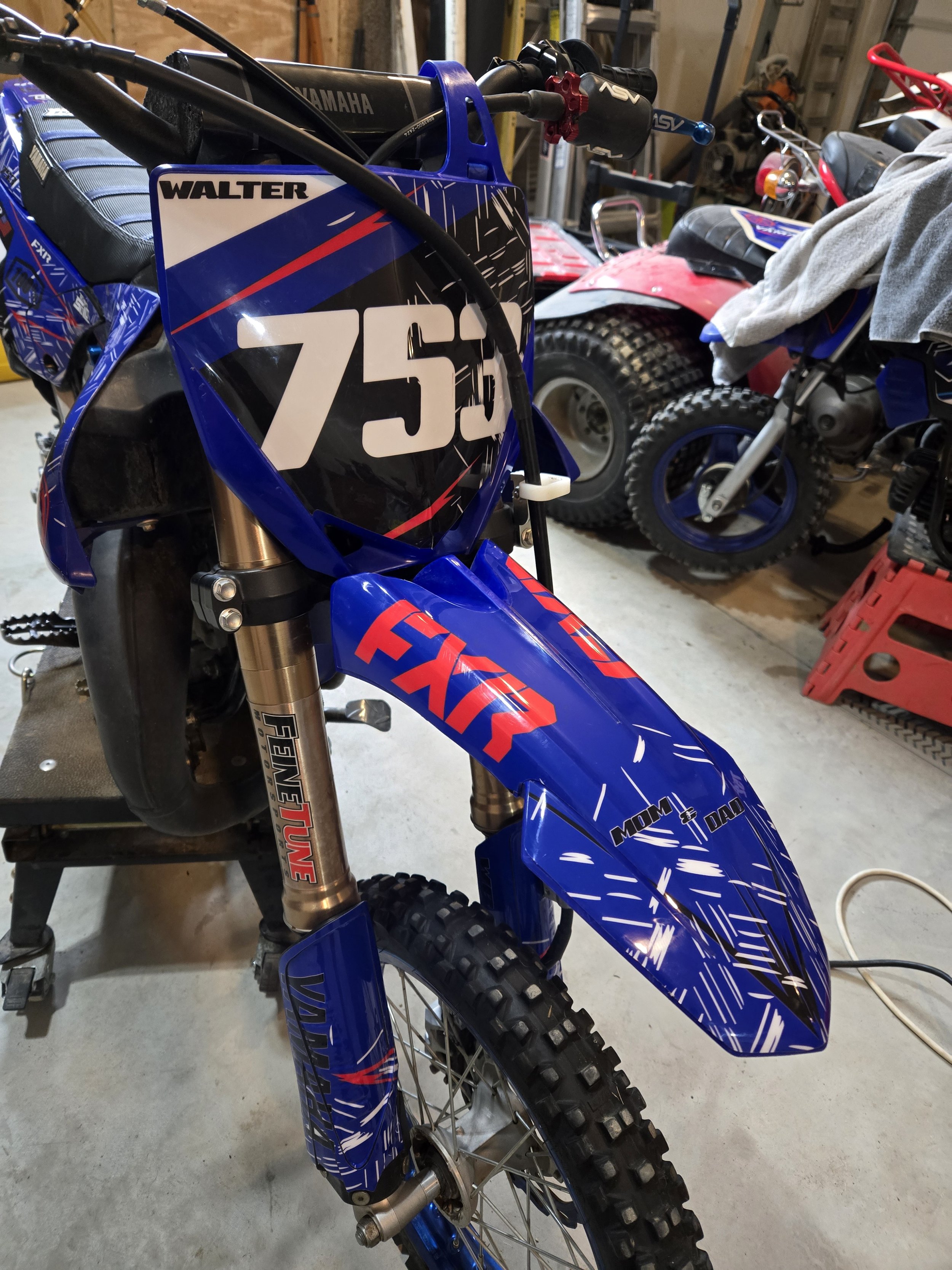 Blue Yamaha dirt bike with the number 750 on the front, featuring Fox racing suspension and knobby tires, parked indoors in a garage with other bikes and tools visible in the background.