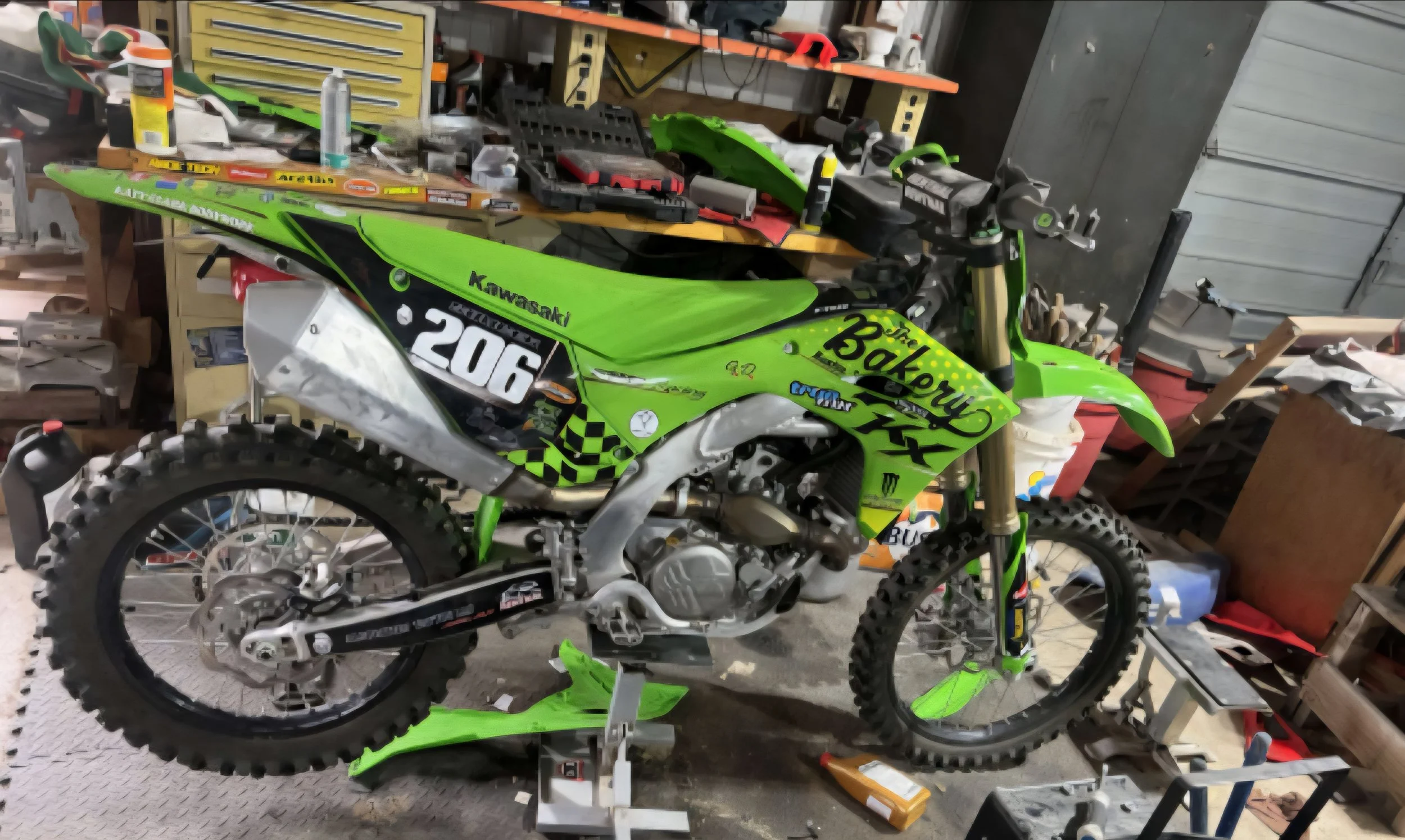 A green Kawasaki dirt bike is in a workshop, surrounded by tools and equipment on a workbench and shelves.