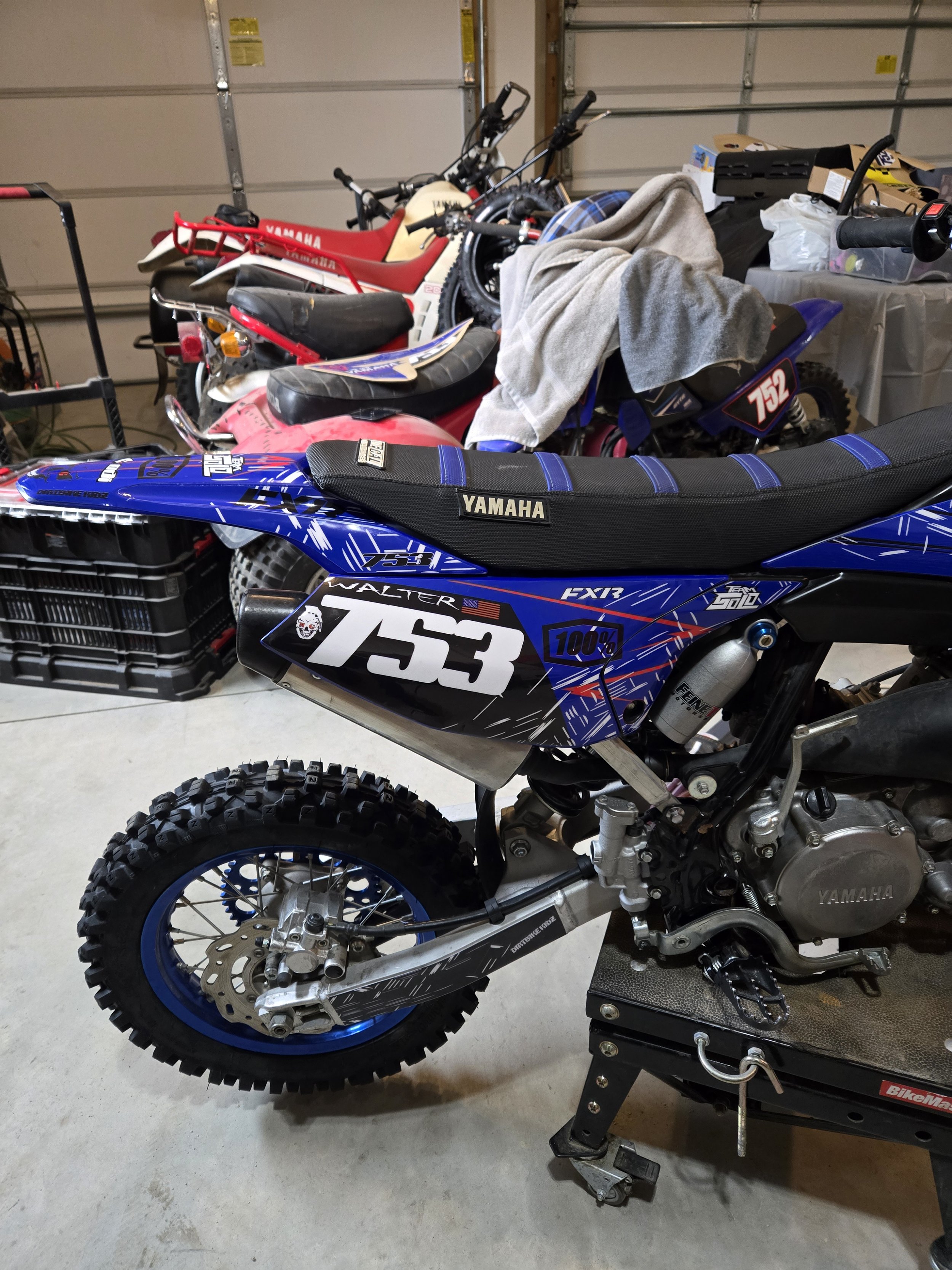 A Yamaha dirt bike in a garage with several other dirt bikes in the background. The bike features a blue and black color scheme, racing number 753, and off-road tires.