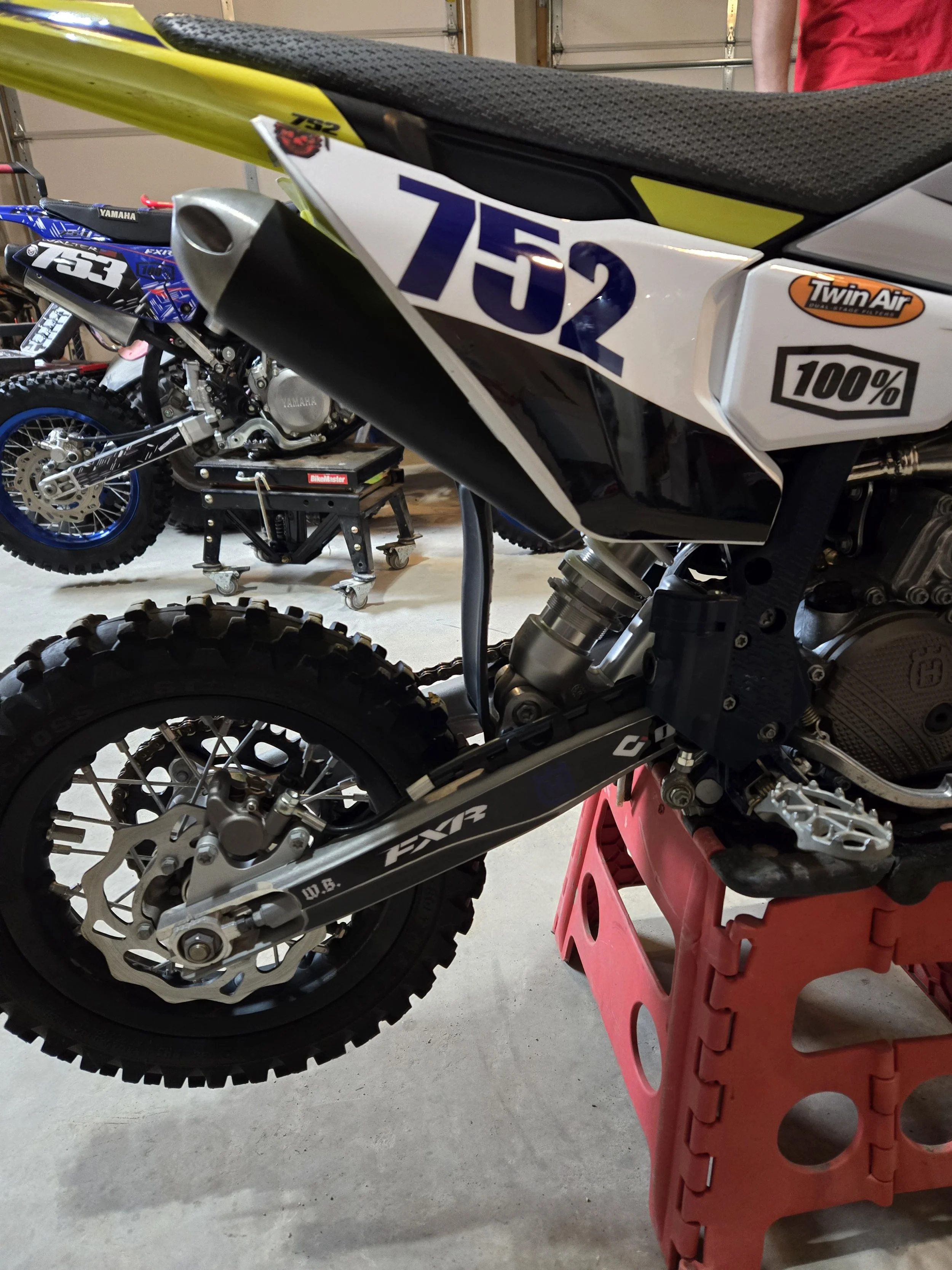 Close-up of a dirt bike with the number 752 on the side, riding on a red stand in a garage. Behind it are more motorcycles, including one with a blue and black color scheme.
