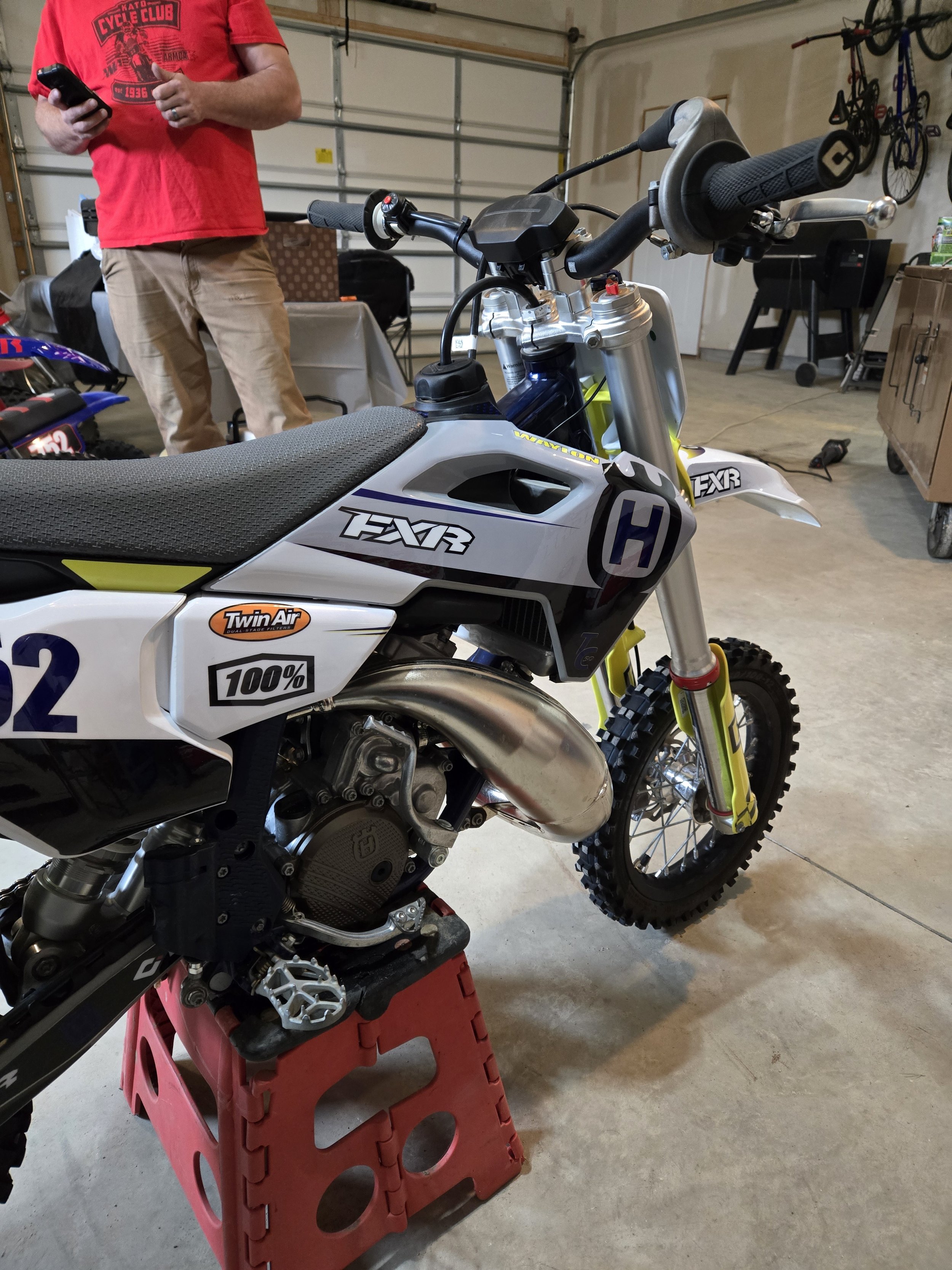 A dirt bike is in a garage, with a person standing nearby holding a phone. The bike has a gray and white body with black, yellow, and blue accents, and is mounted on a red stand.