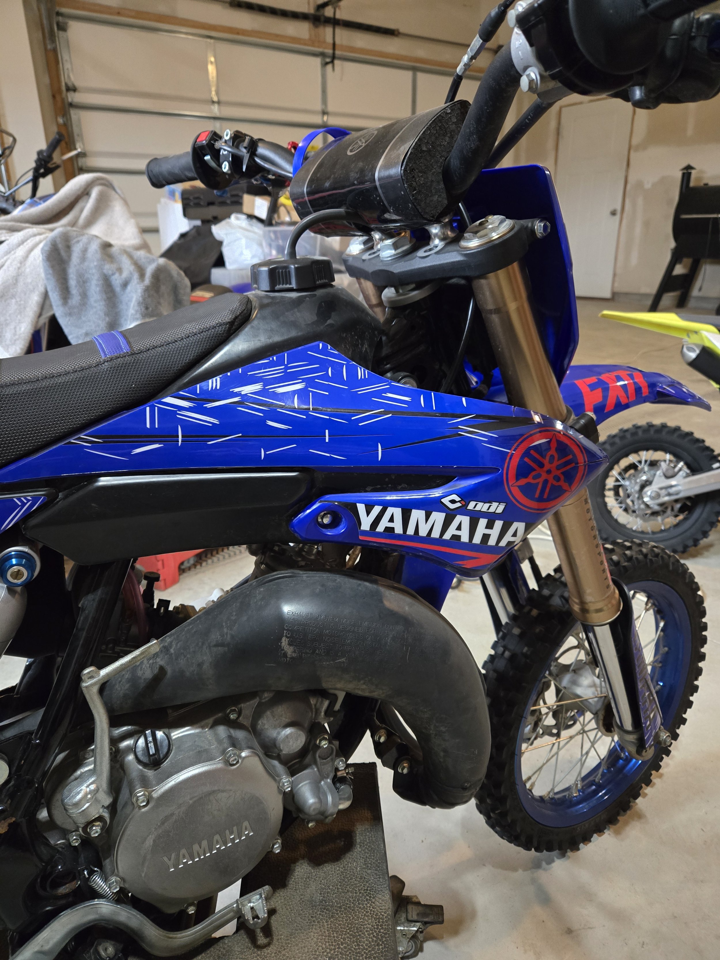 Close-up view of a Yamaha dirt bike with a blue and black color scheme, positioned in a garage with other bikes and tools visible in the background.