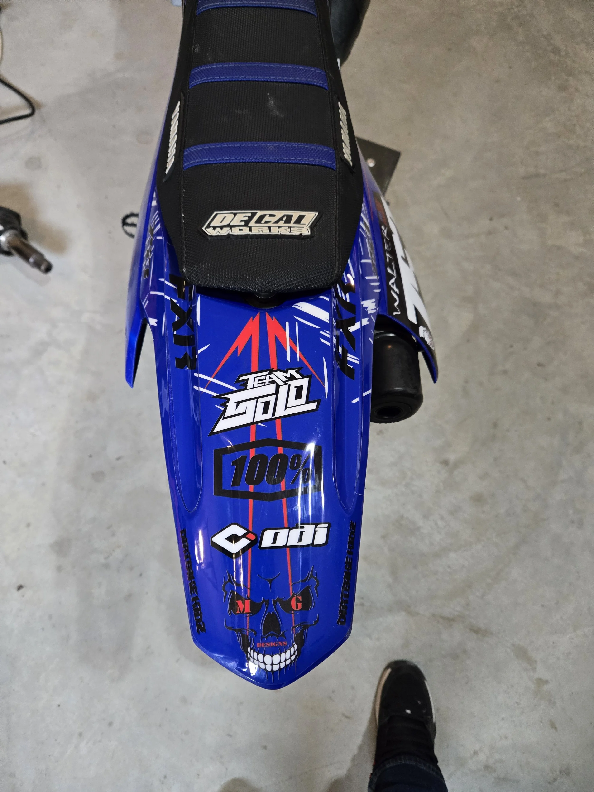 Top view of a blue motocross dirt bike with various stickers, including a skull, 'TEAM SOUL', '100%', 'DGI', and 'Decal Works'; a black and blue seat with a sticker.