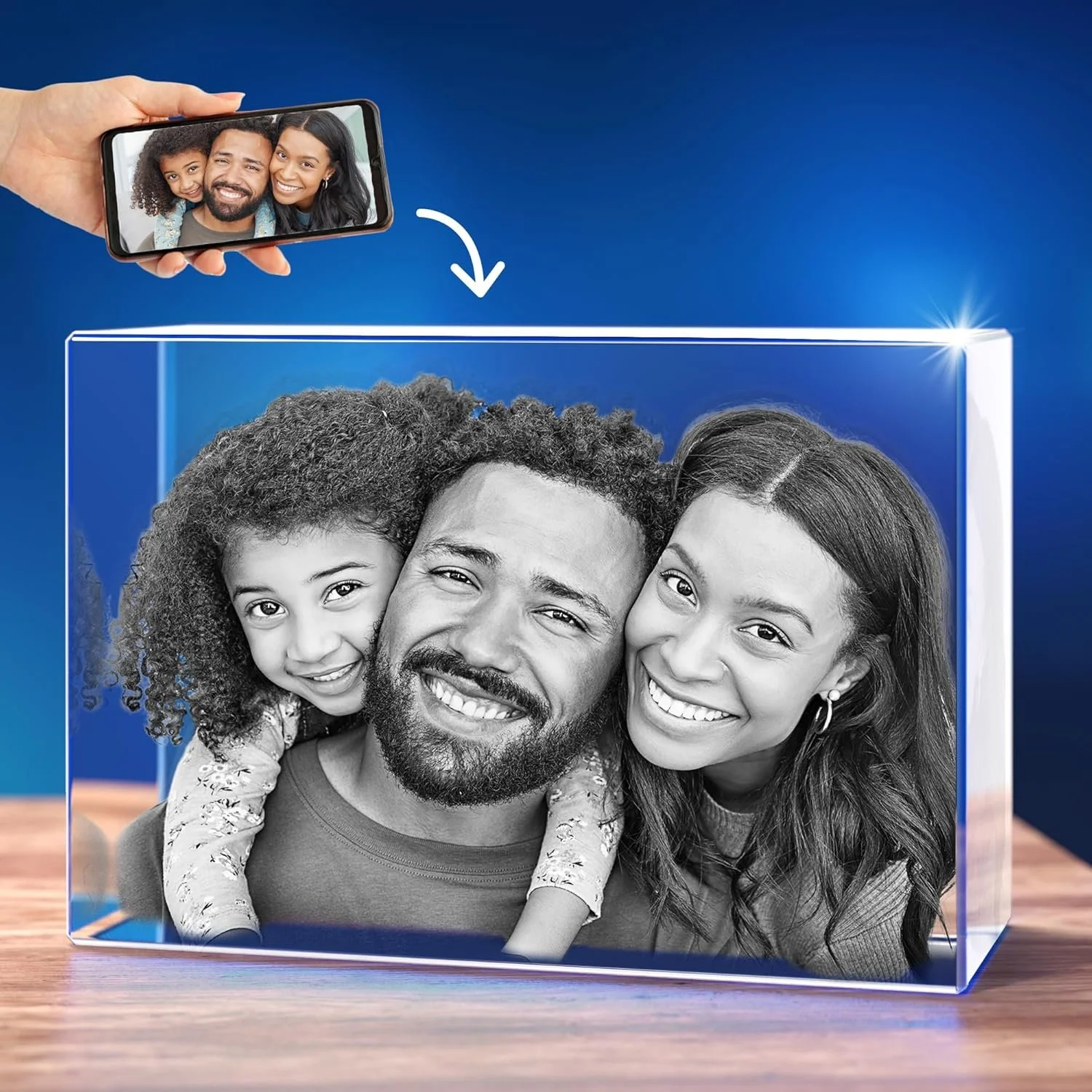 A clear 3D acrylic block containing a black and white photo of a happy family of three, including a man with a beard, a woman with long hair, and their young daughter. A hand holds a smartphone above the block, showing a color photo of the same family.
