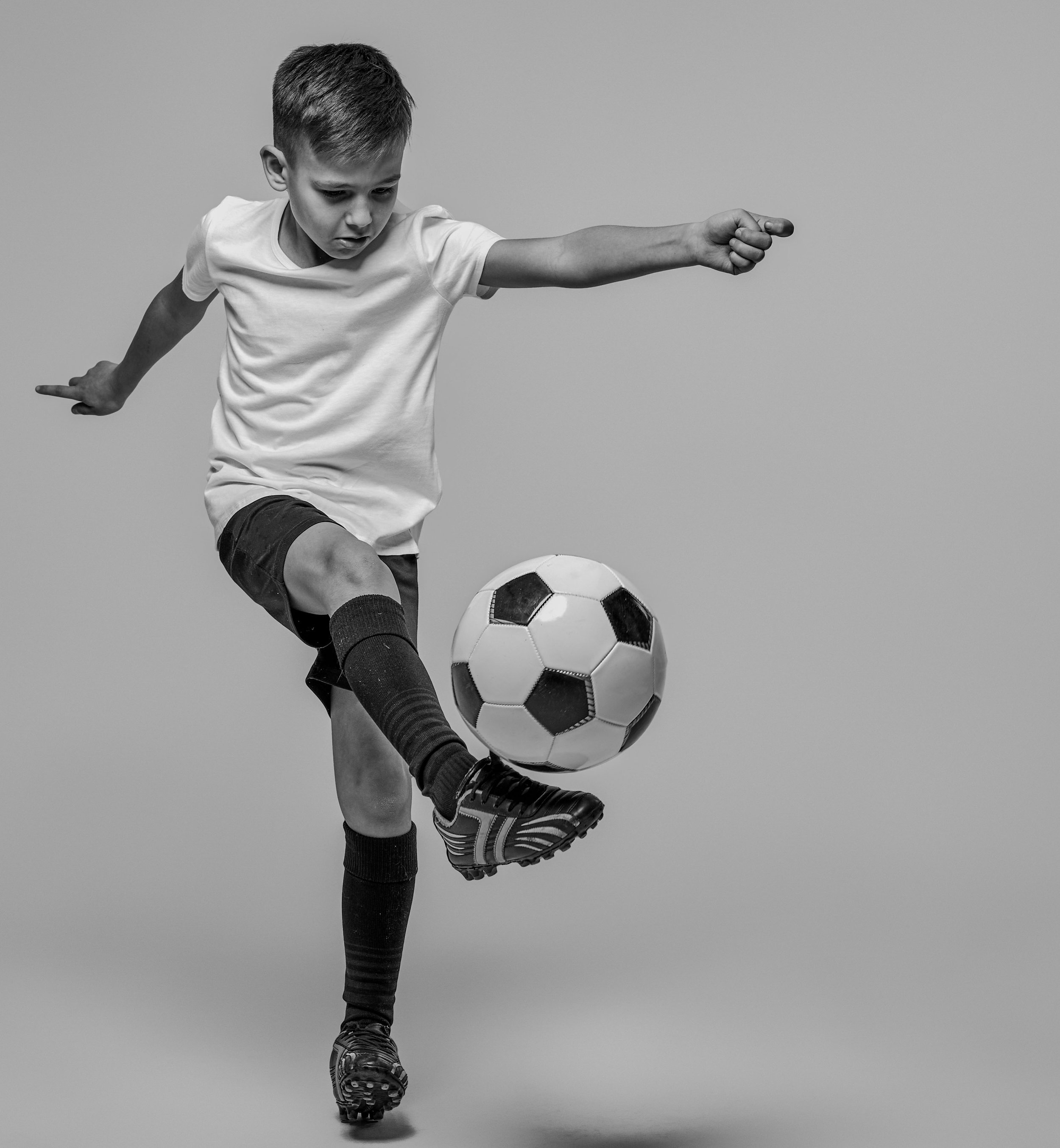 A young boy wearing a t-shirt, shorts, and soccer shoes is kicking a soccer ball in a studio with a plain background.