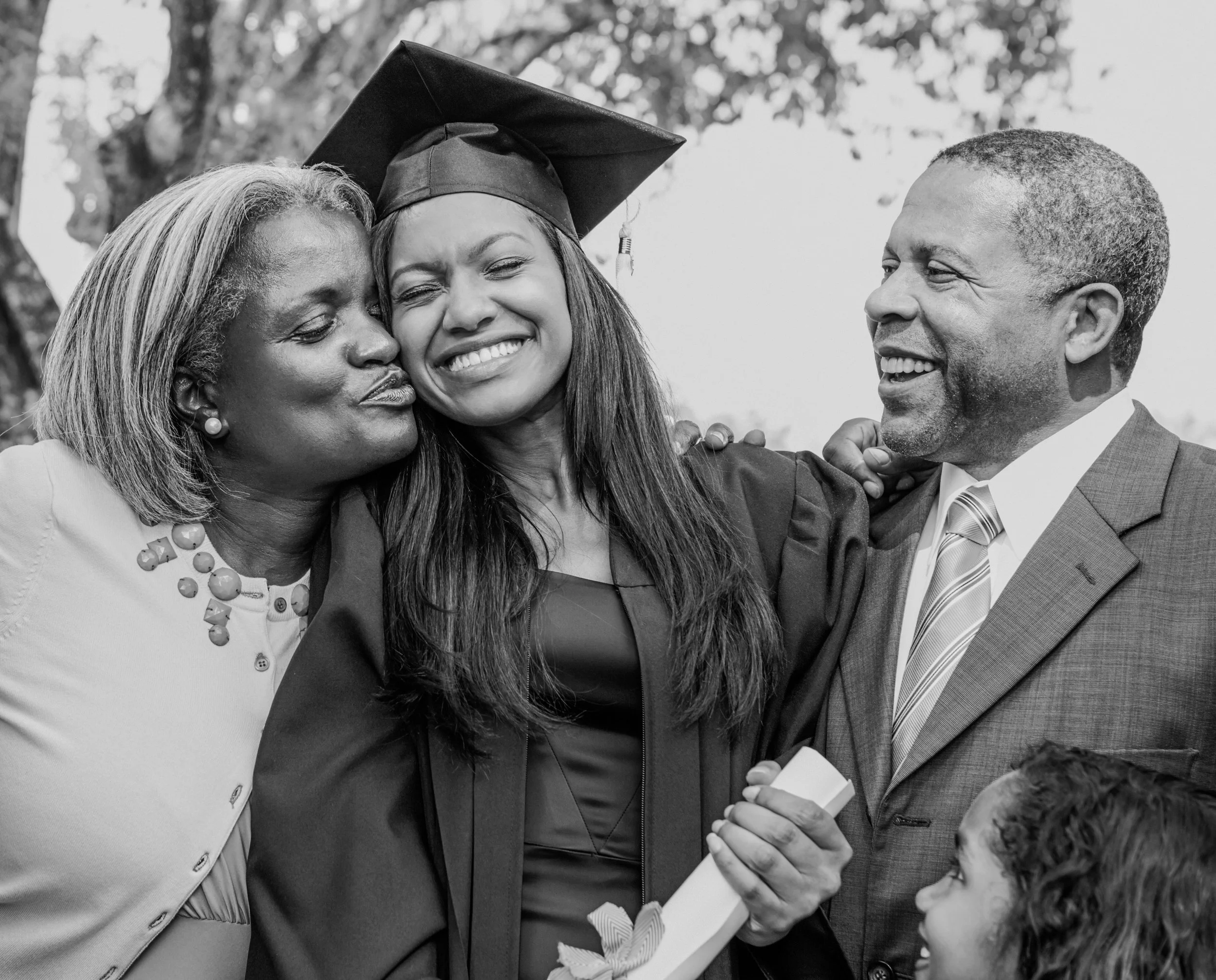 A young woman in graduation gown and cap hugging her parents, who are smiling with her during her graduation celebration.