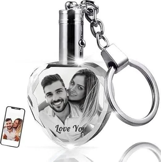 A heart-shaped lock with a photo of a smiling couple and the words "Love You" inside it. The lock is attached to a keychain and a necklace chain, with a smartphone showing the same photo of the couple.
