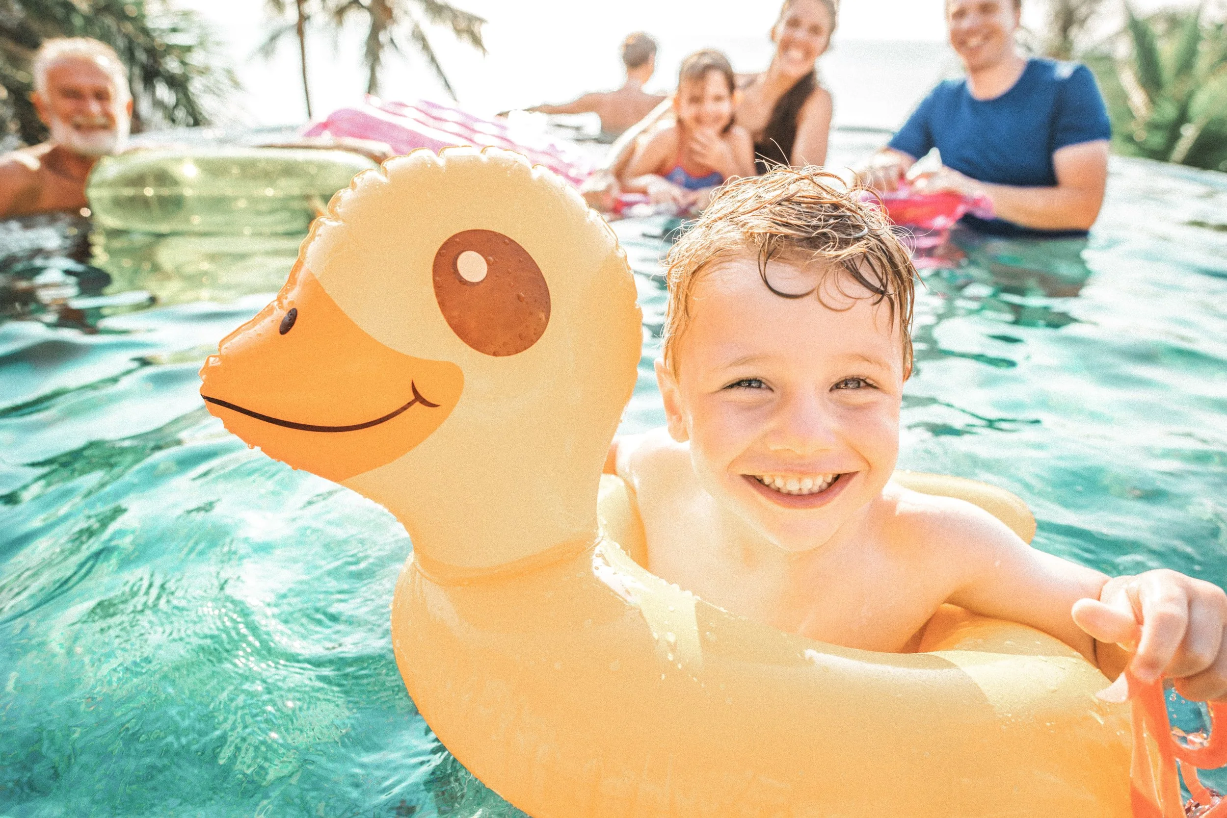 A young boy with a big smile in a swimming pool, holding a yellow duck-shaped pool float, with a group of smiling people in the background.