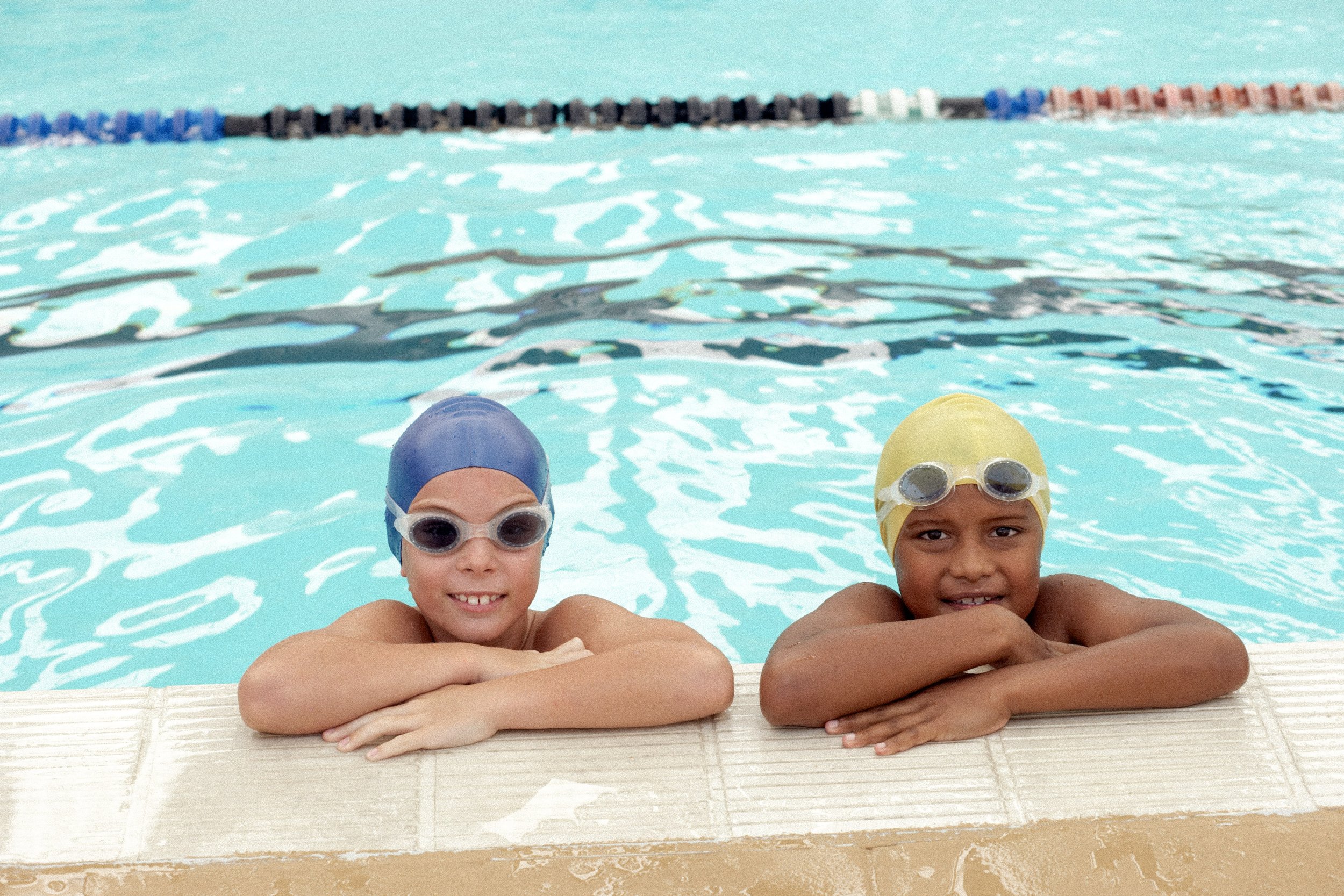 Two young children wearing swim caps and goggles, leaning on the edge of a swimming pool, smiling.