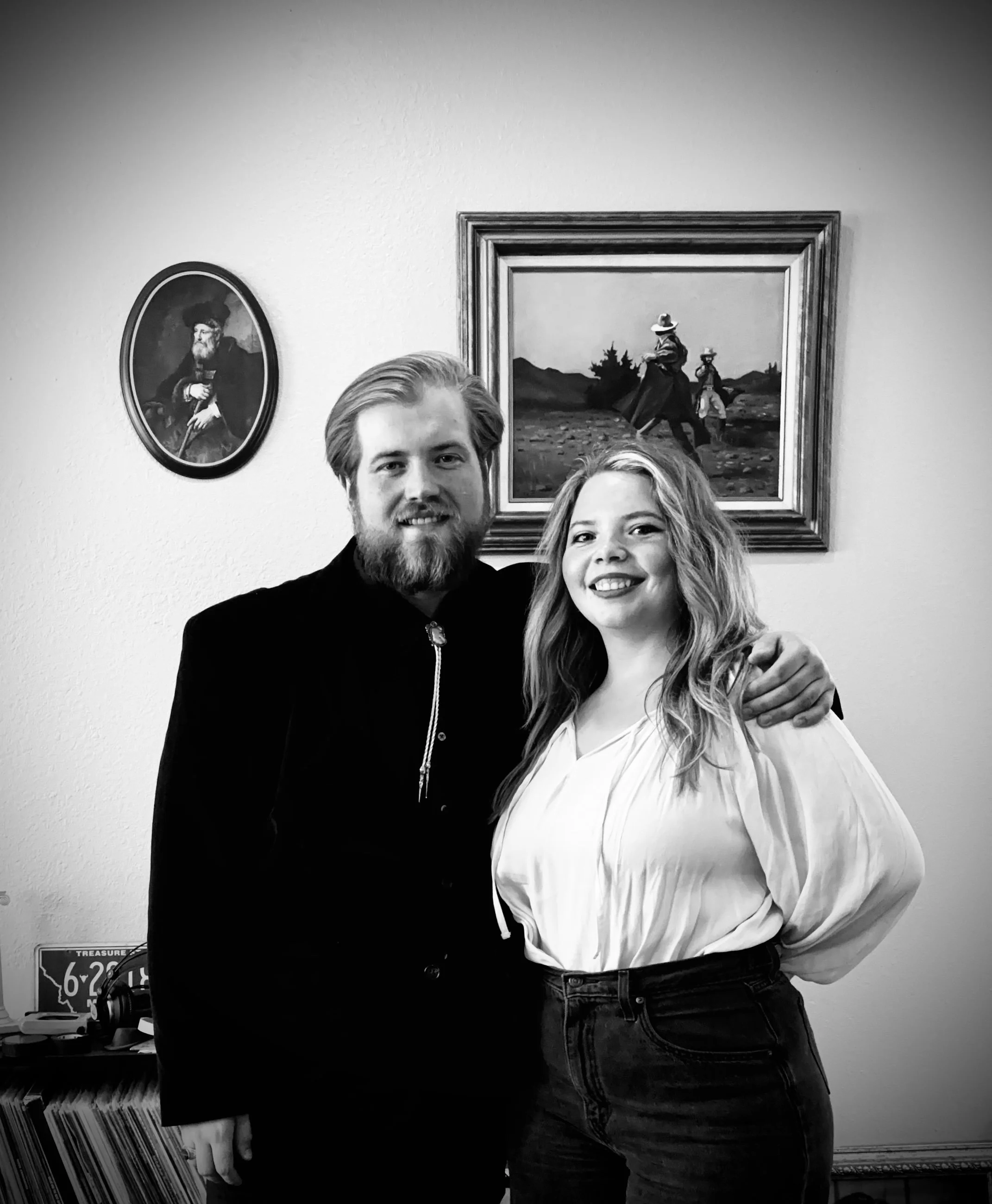 A black and white photo of a man with a beard and a woman with curly hair smiling and posing together in front of a wall adorned with framed paintings. The man has his arm around the woman, and they are both facing the camera.