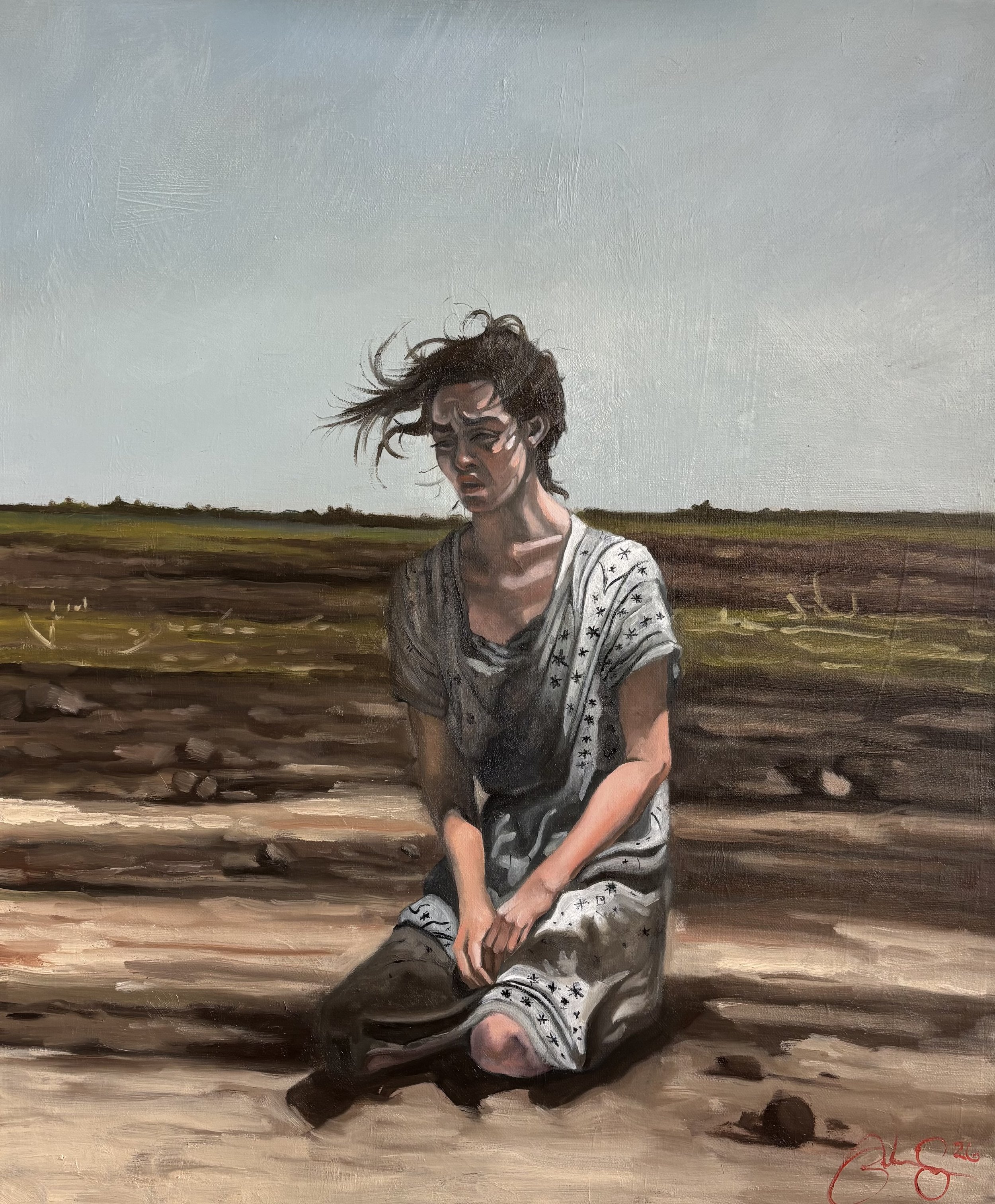 Young woman with short hair and closed eyes, sitting outdoors on a dirt field, wearing a gray and black star-patterned shirt. Her hair is being blown by the wind, and she appears to be upset or distressed.