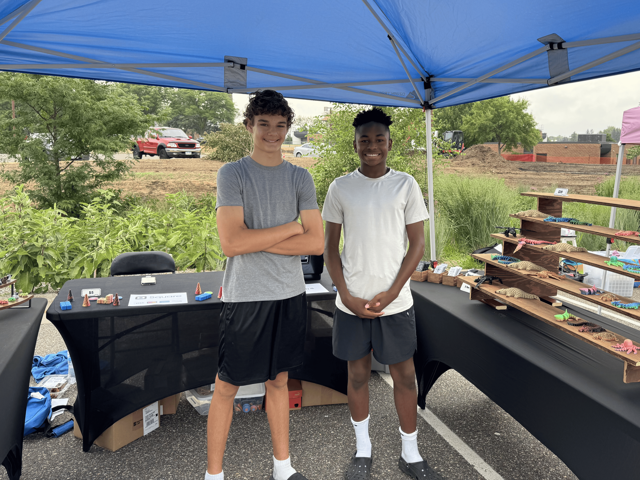 Two young boys standing under a blue canopy at an outdoor market stall, with jewelry and trinkets displayed on tables.