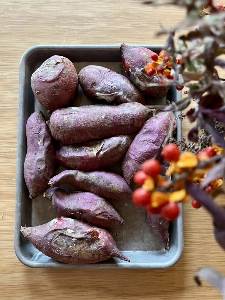 A metal tray on a wooden surface containing multiple purple sweet potatoes. In the foreground, there is a blurry branch with red berries and small yellow flowers.