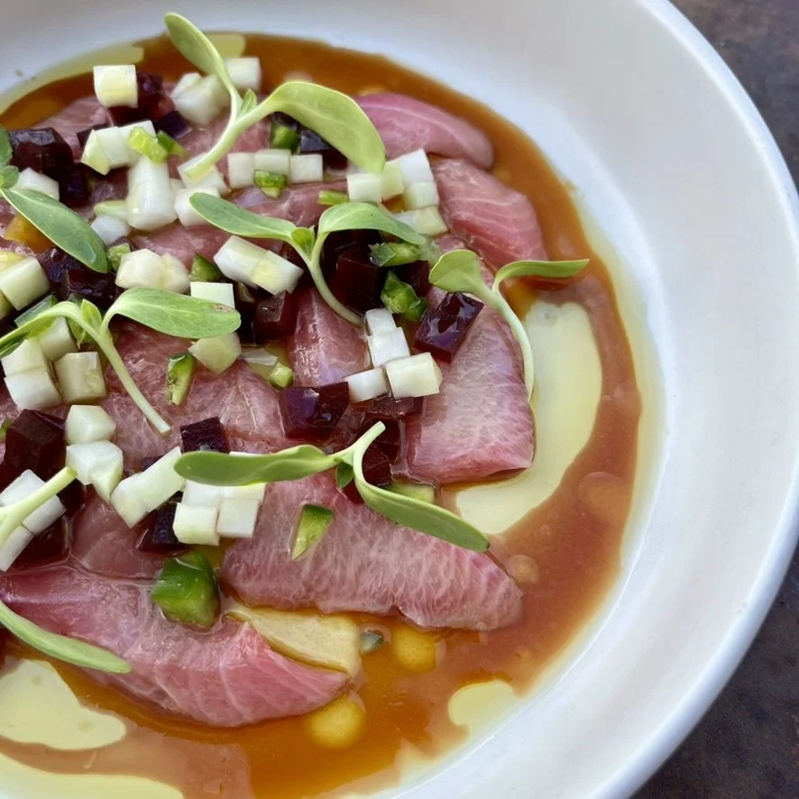 Thinly sliced raw beef garnished with chopped green onions, microgreens, and diced purple and white vegetables, served with sauce in a white bowl.