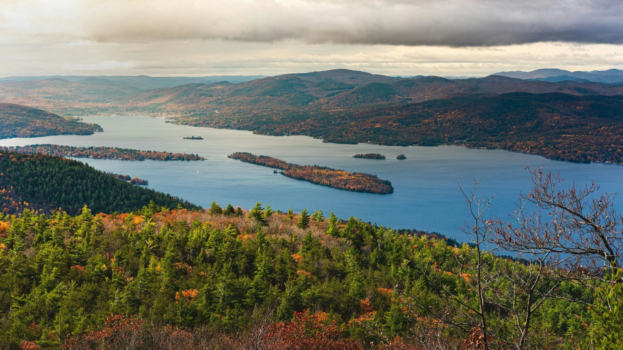 Aerial view of a large lake with multiple islands surrounded by forested hills and mountains, showing fall foliage with green, orange, and red trees under a cloudy sky.