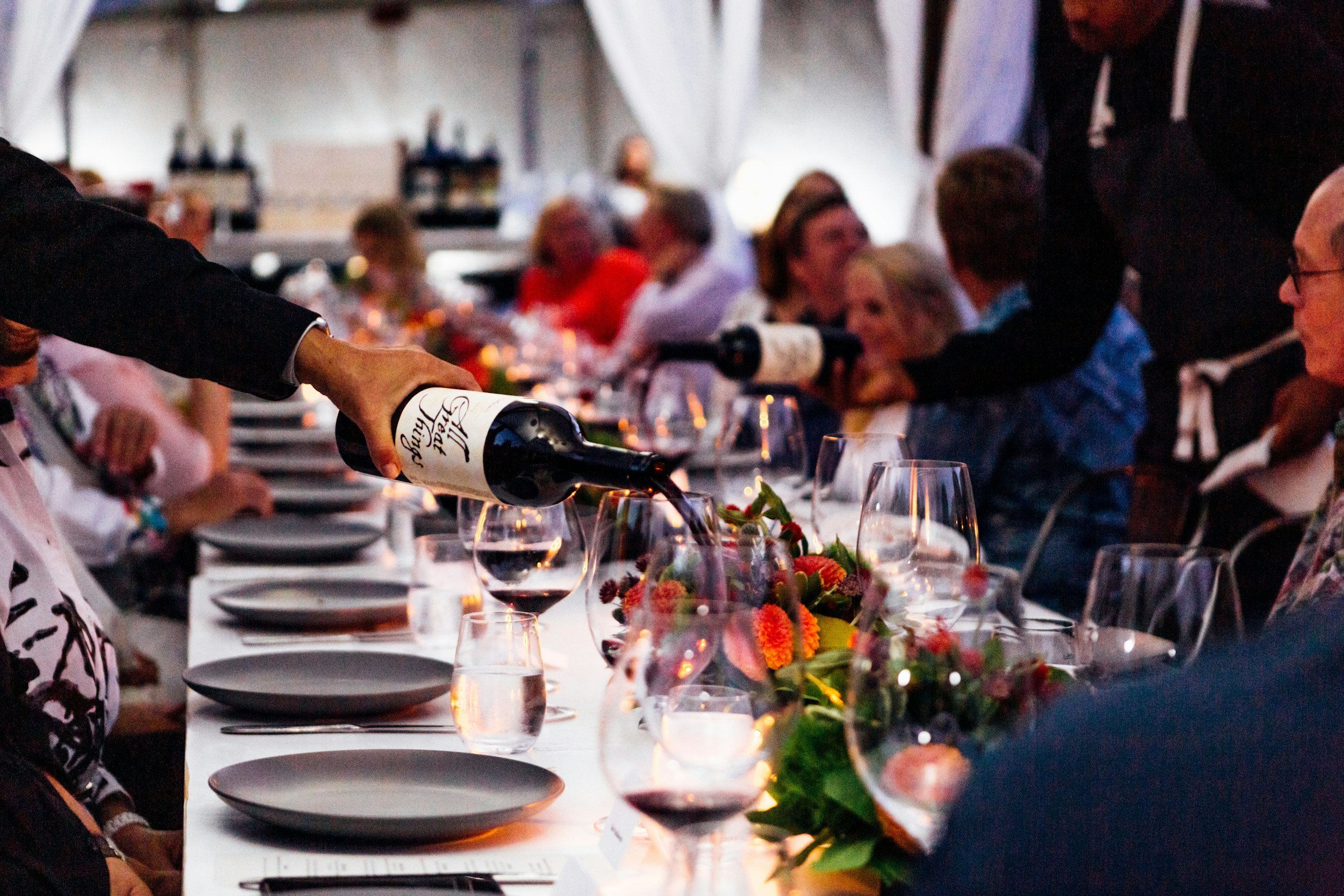 People seated at a long dinner table with wine glasses, floral centerpieces, and a person pouring red wine from a bottle.